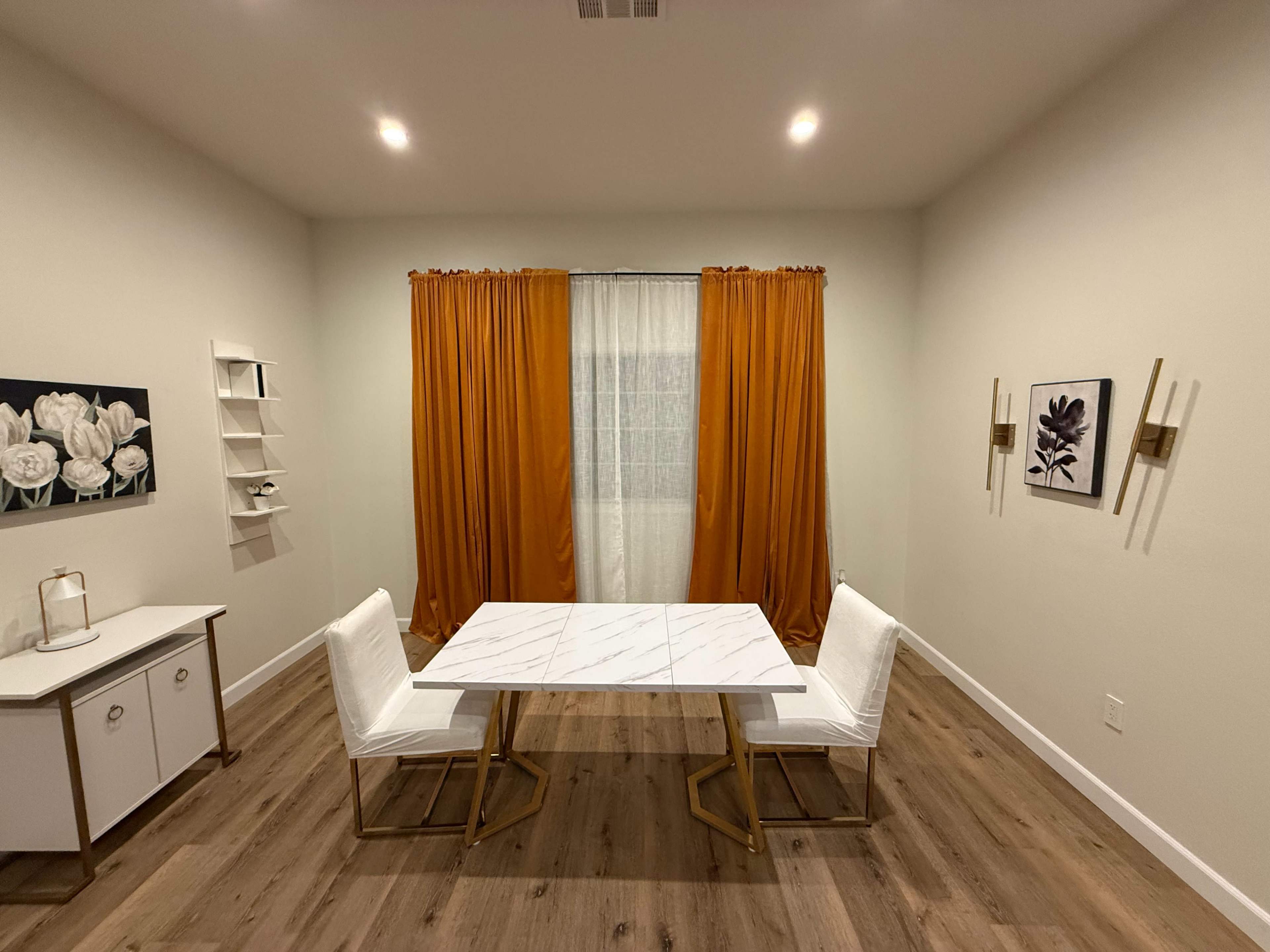 The image shows a modern, minimalist dining area featuring a marble-top table with two white chairs, orange curtains, and minimal wall decor.