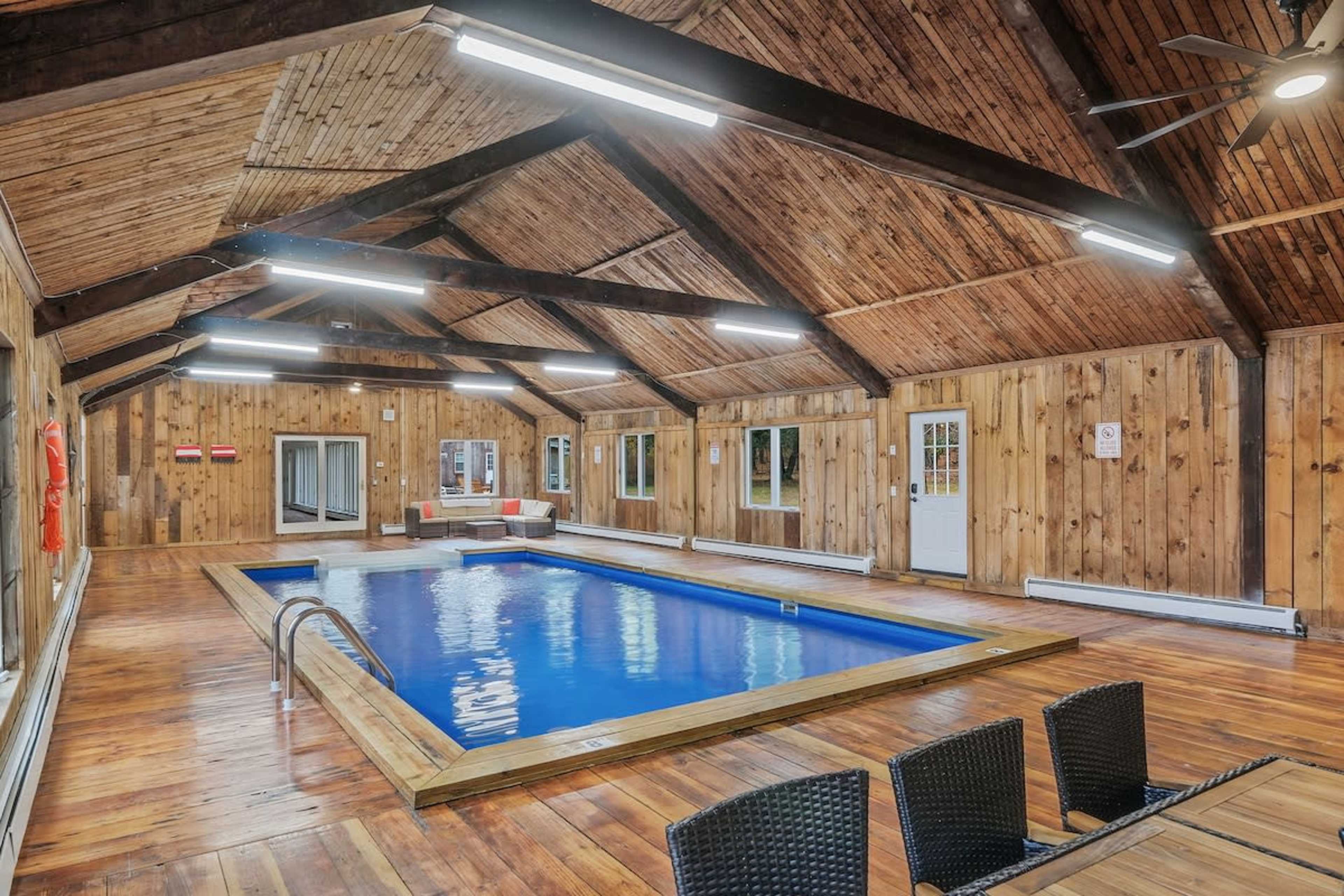 The image shows an indoor wooden structure with a swimming pool at the center, surrounded by seating and large windows.
