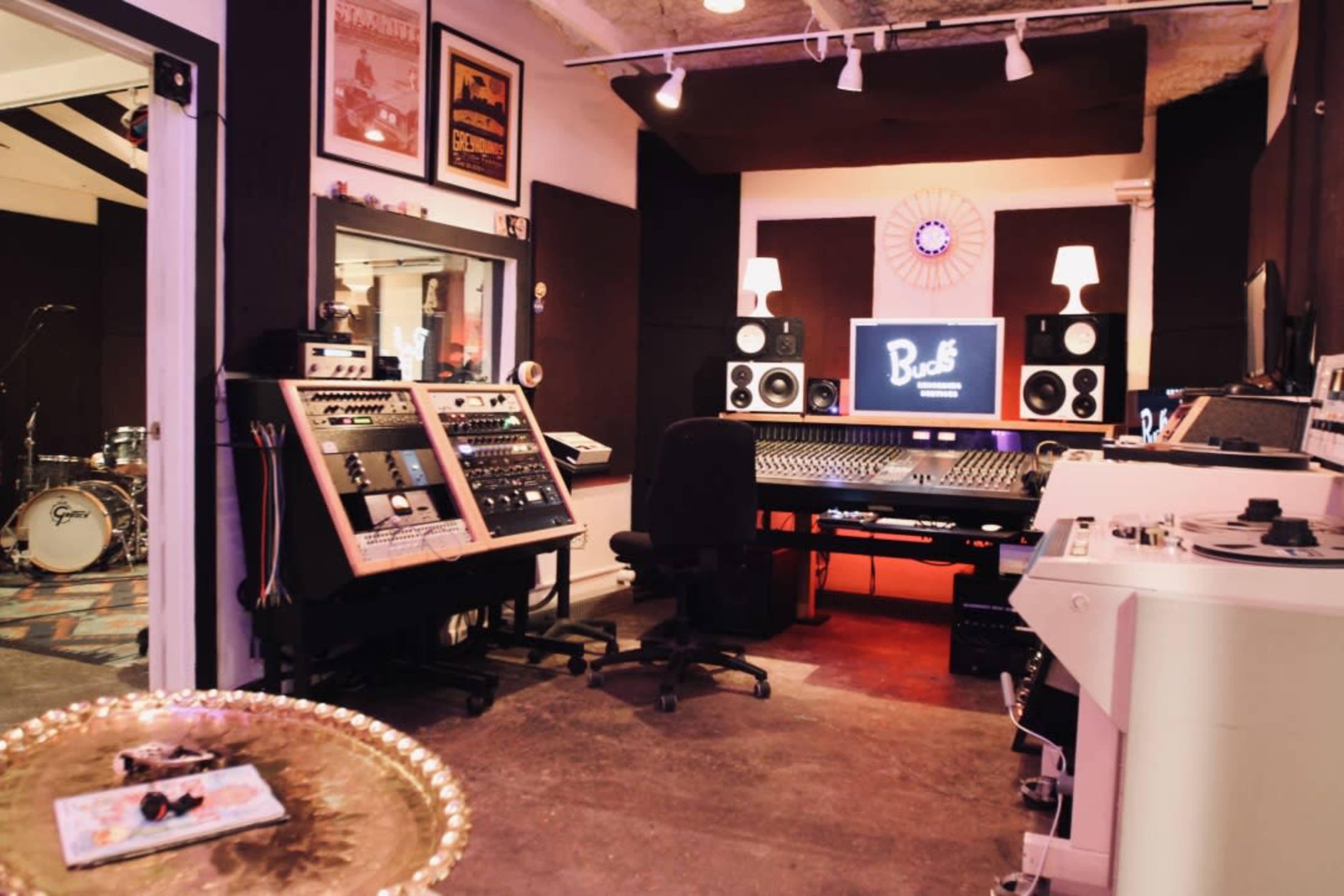 The image shows a music recording studio with a control desk, audio equipment, and soundproofing panels on the walls.