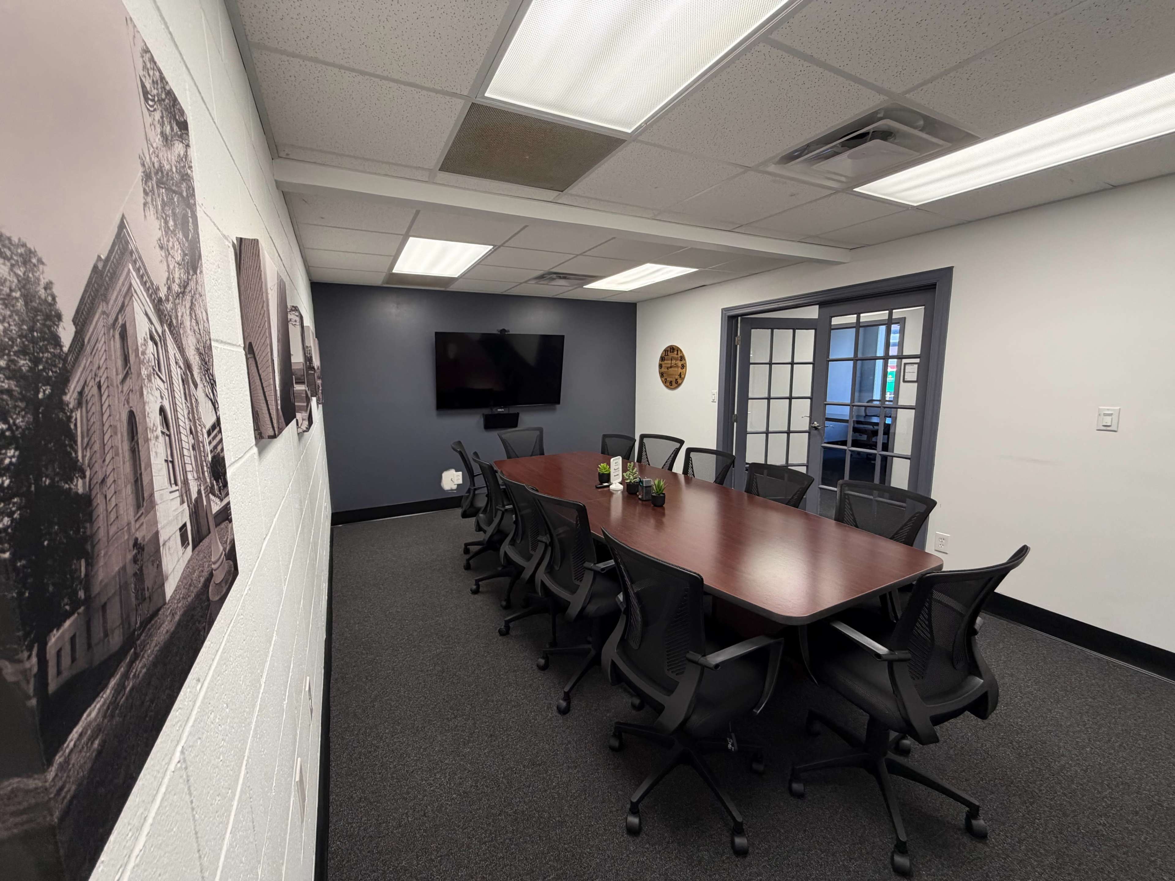 A conference room features a long wooden table surrounded by black swivel chairs, with a television mounted on the wall and large windows offering a view of the outside.