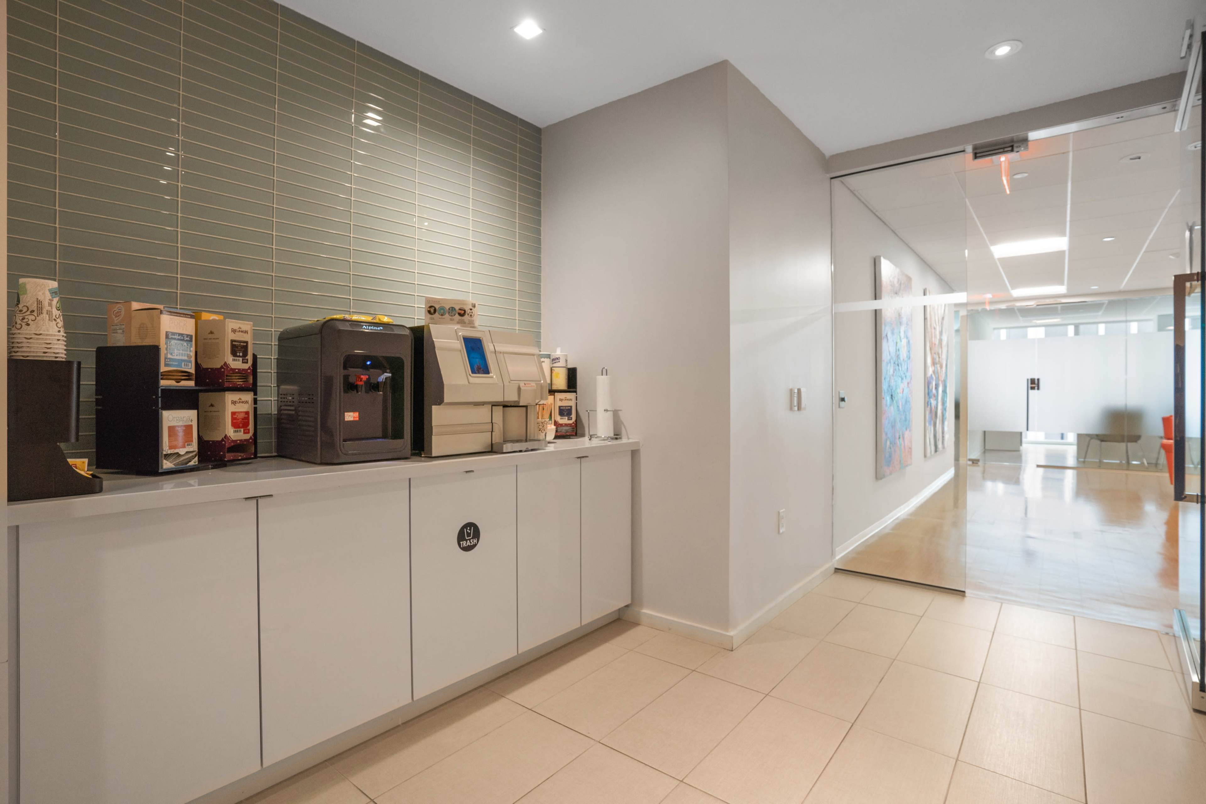 A modern break room with a coffee machine and a counter against a tiled wall, leading to an open corridor with artwork.