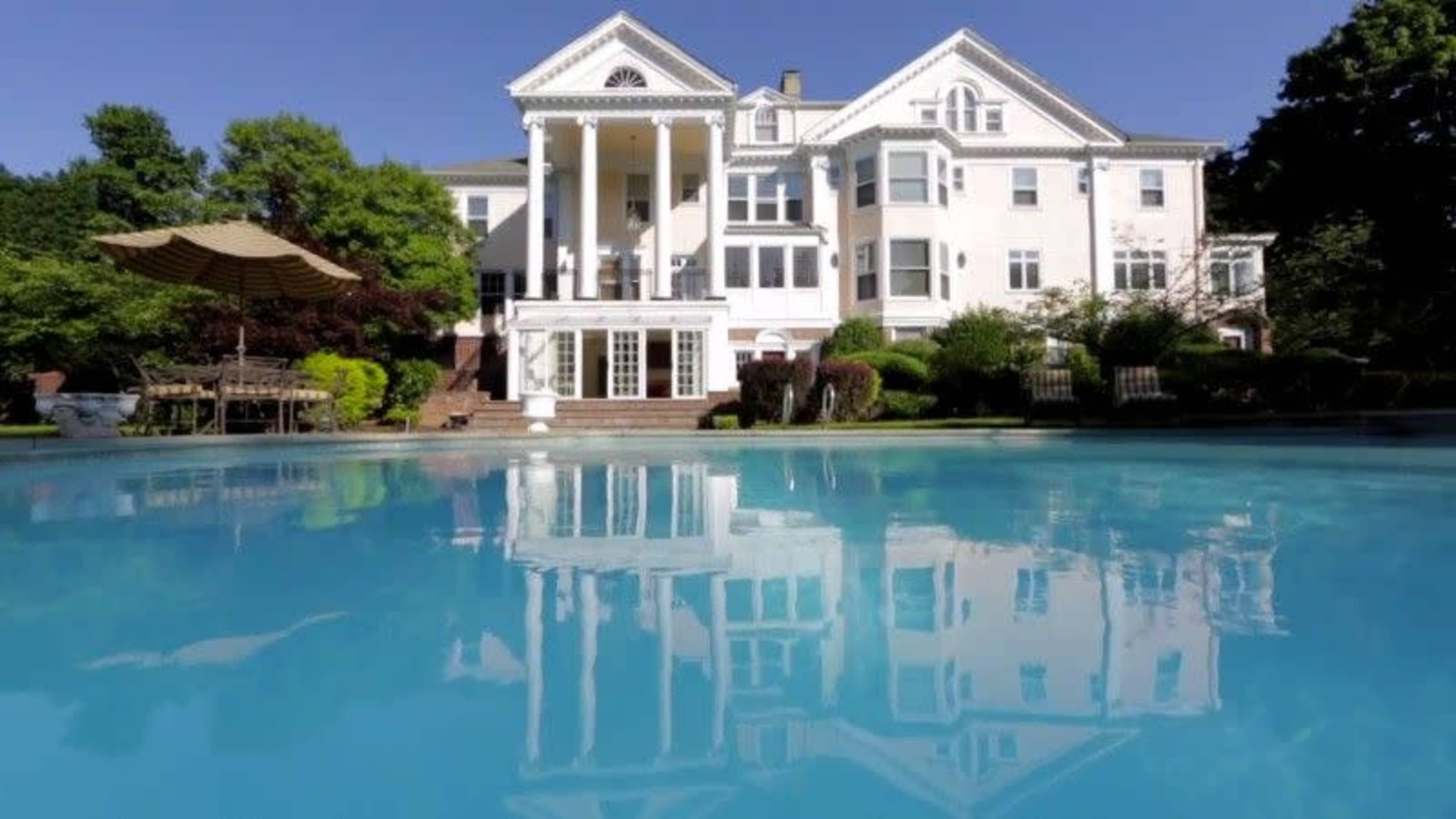 A large, white mansion with columns is reflected in a clear blue swimming pool surrounded by greenery.