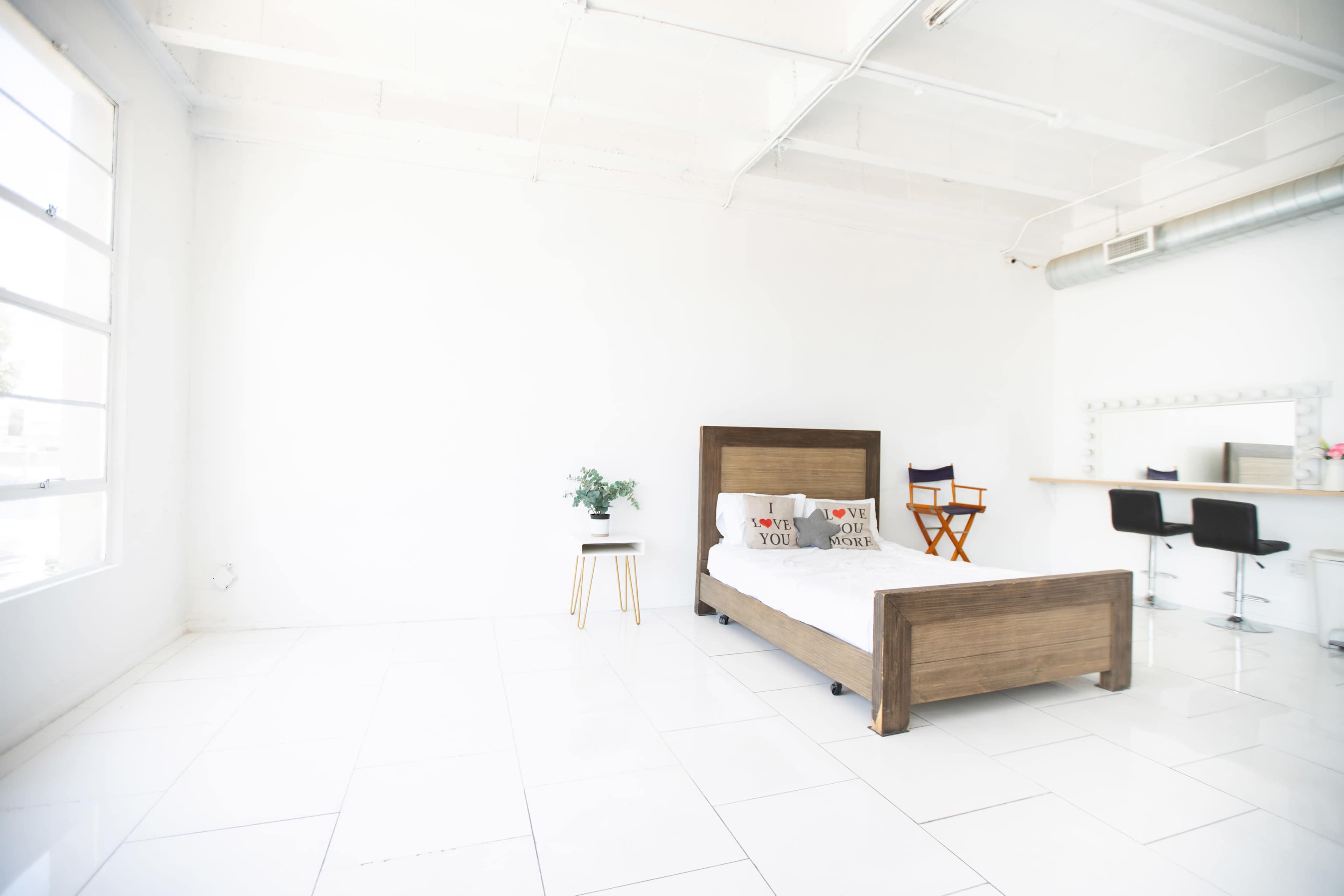 The image shows a minimalist bedroom with a wooden bed, a small plant on a side table, and a bar area with two stools against a white wall.