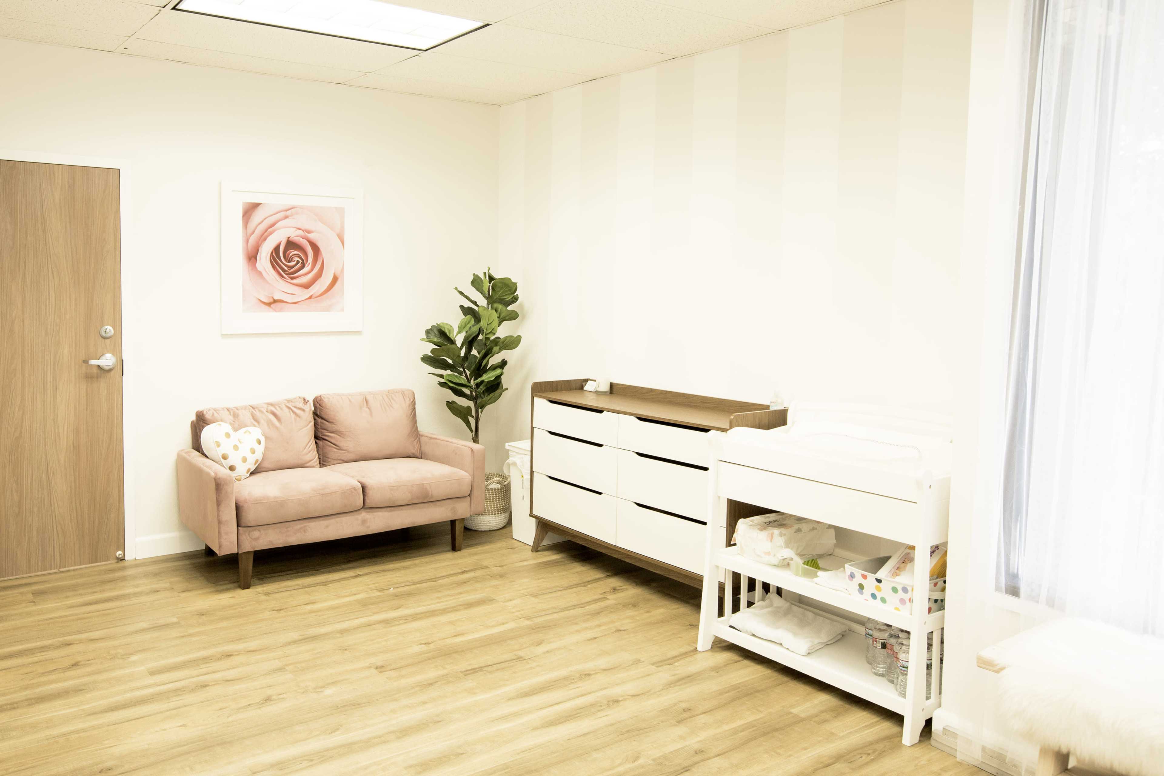 The image shows a bright room with wooden flooring, featuring a pink couch, a dresser, a changing table, and a leafy indoor plant against a striped wall.