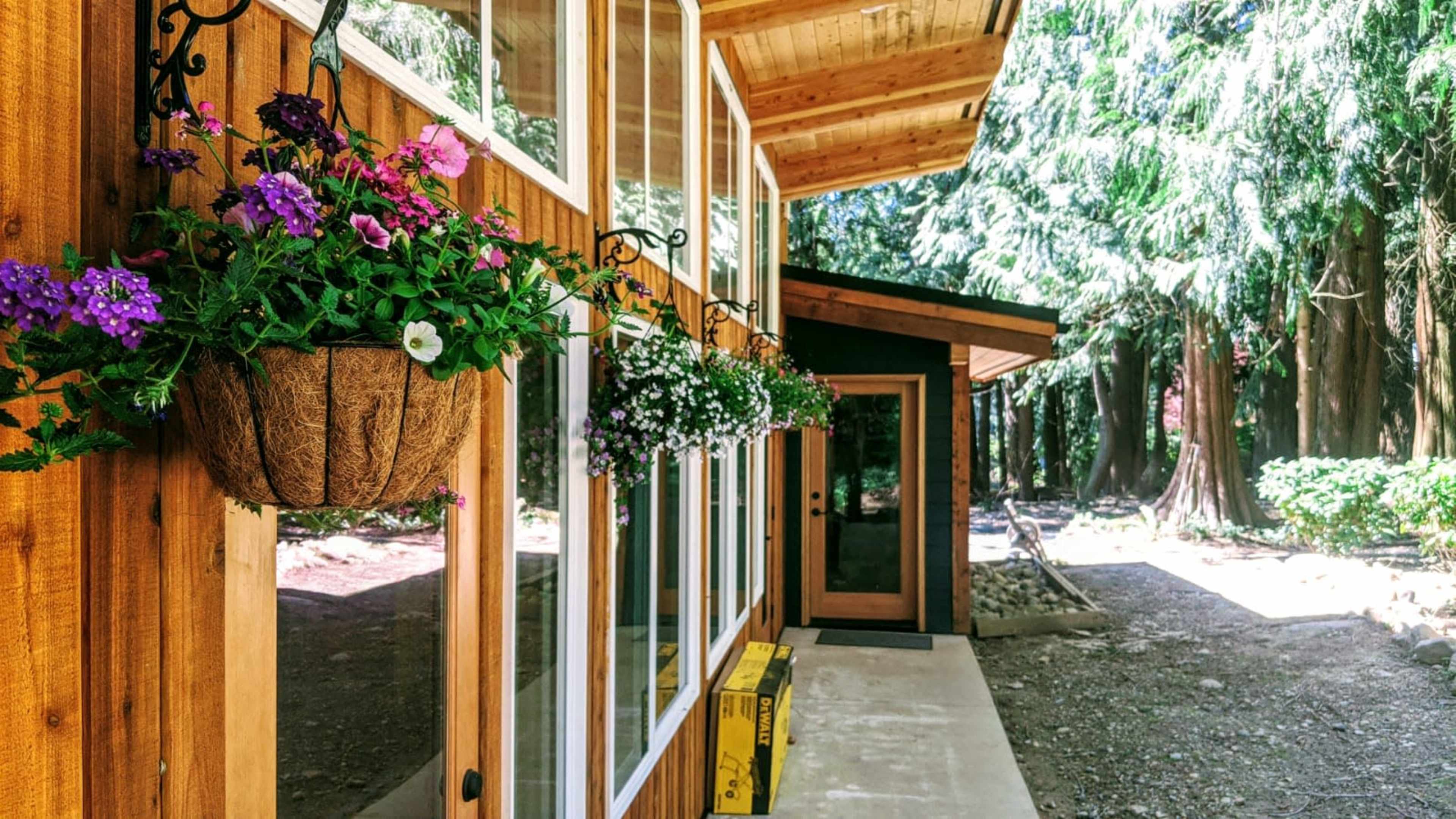 A wooden cabin features large windows adorned with hanging flower baskets in a wooded setting.