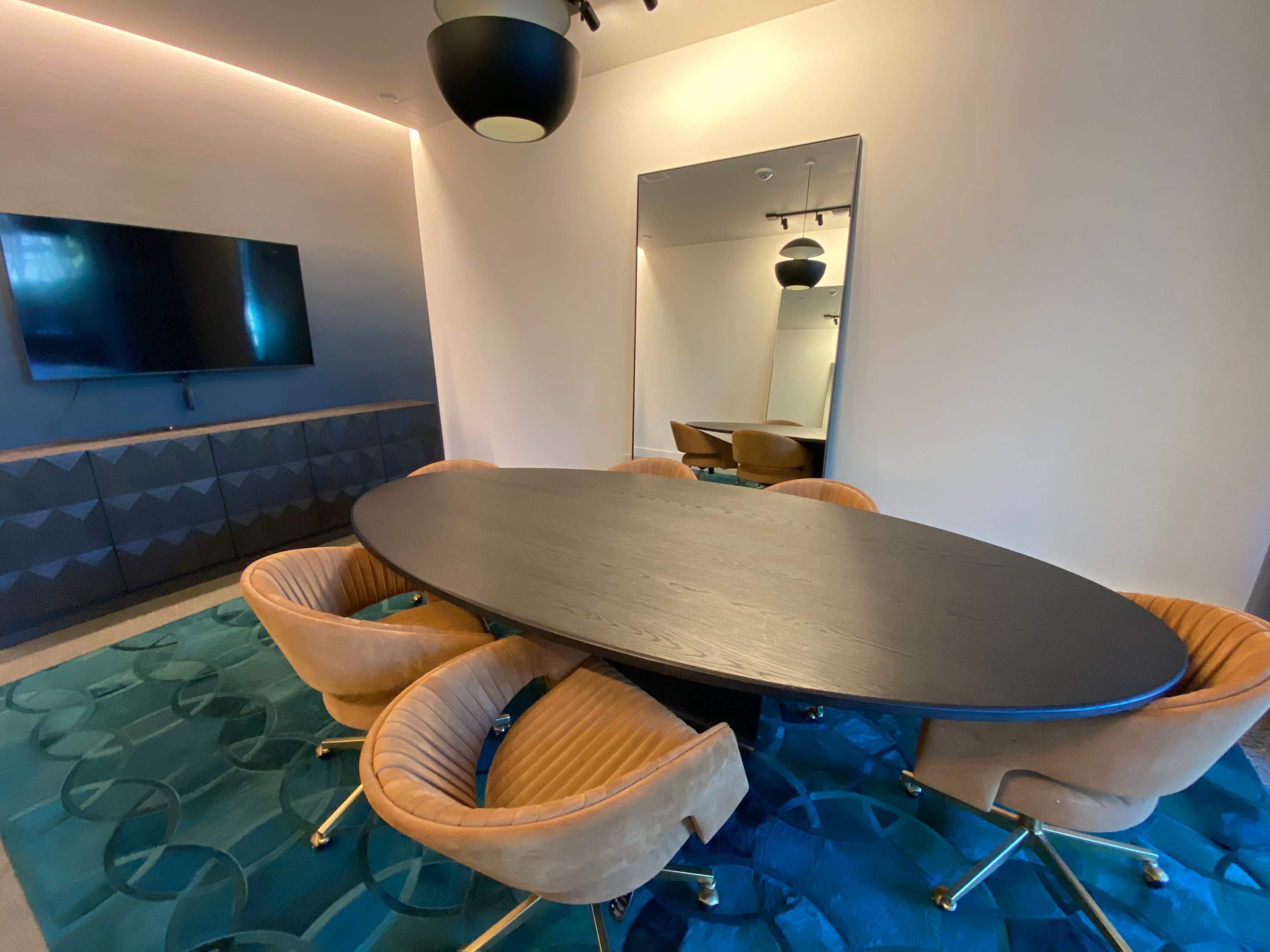 A modern conference room with an oval black table surrounded by five upholstered chairs, a wall-mounted television, and a large mirror.