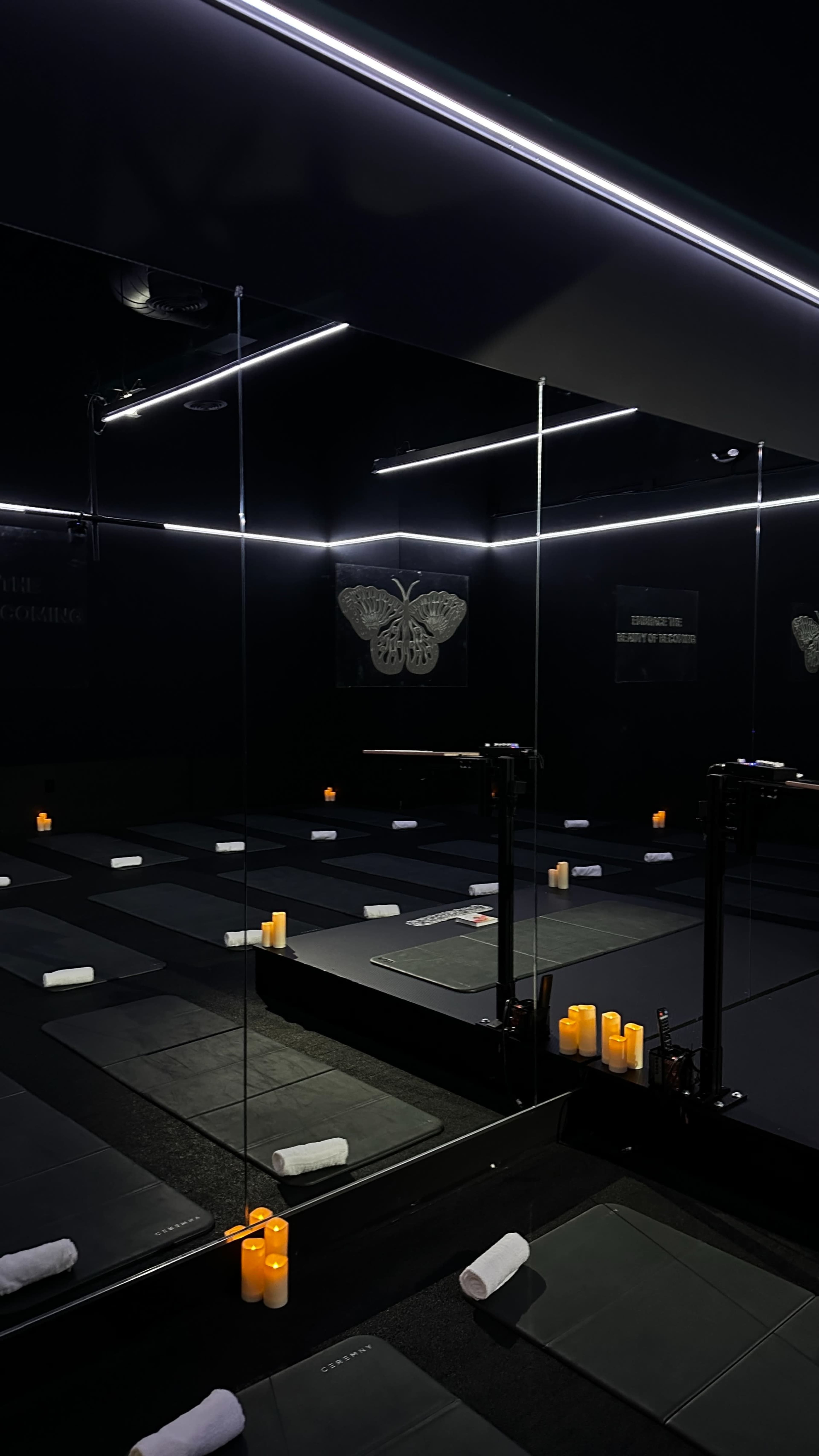 A dimly lit yoga studio features black mats arranged on the floor, surrounded by candles and illuminated by overhead lights.
