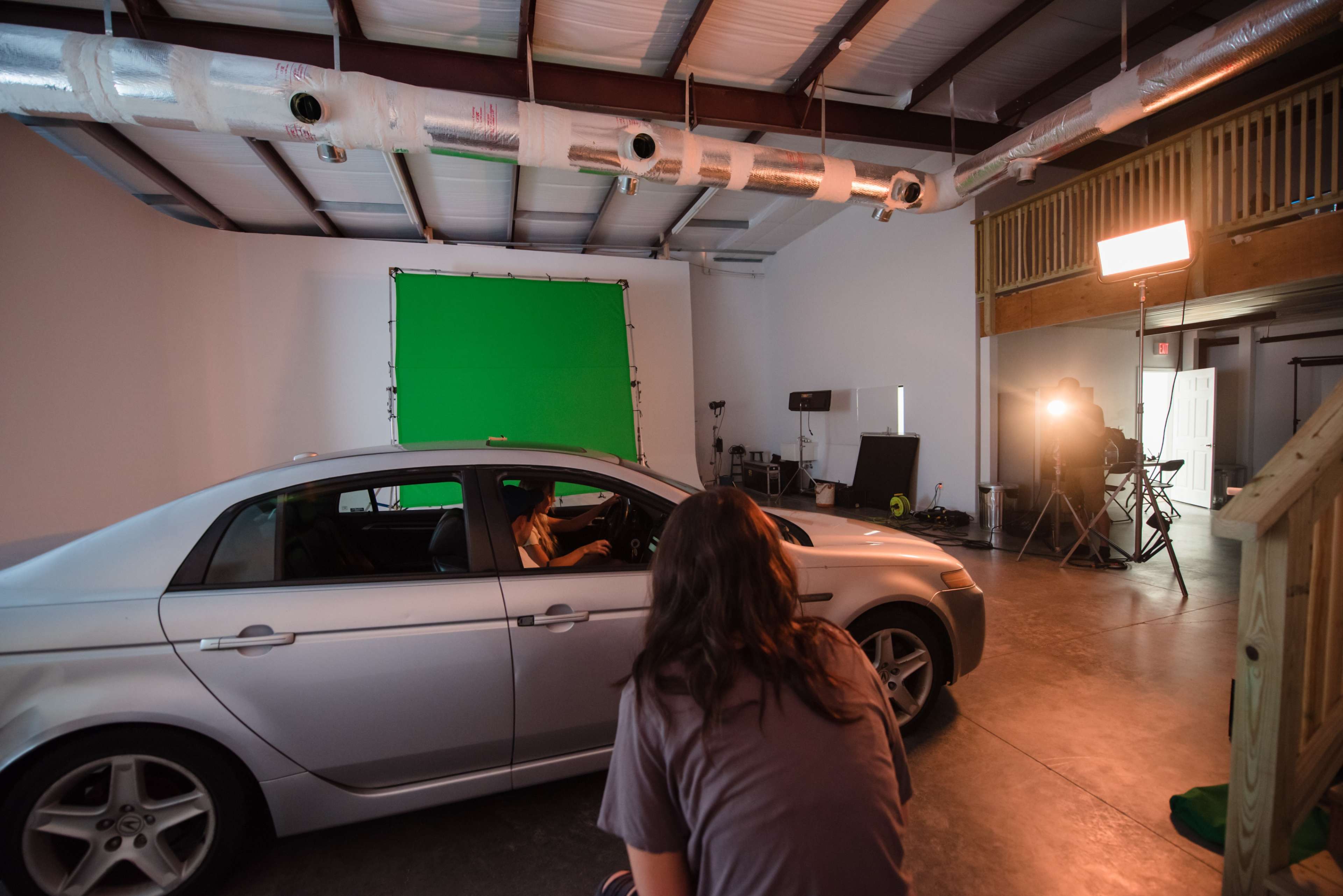 A silver car is positioned in a studio space with a green backdrop and lighting equipment set up for a photoshoot.