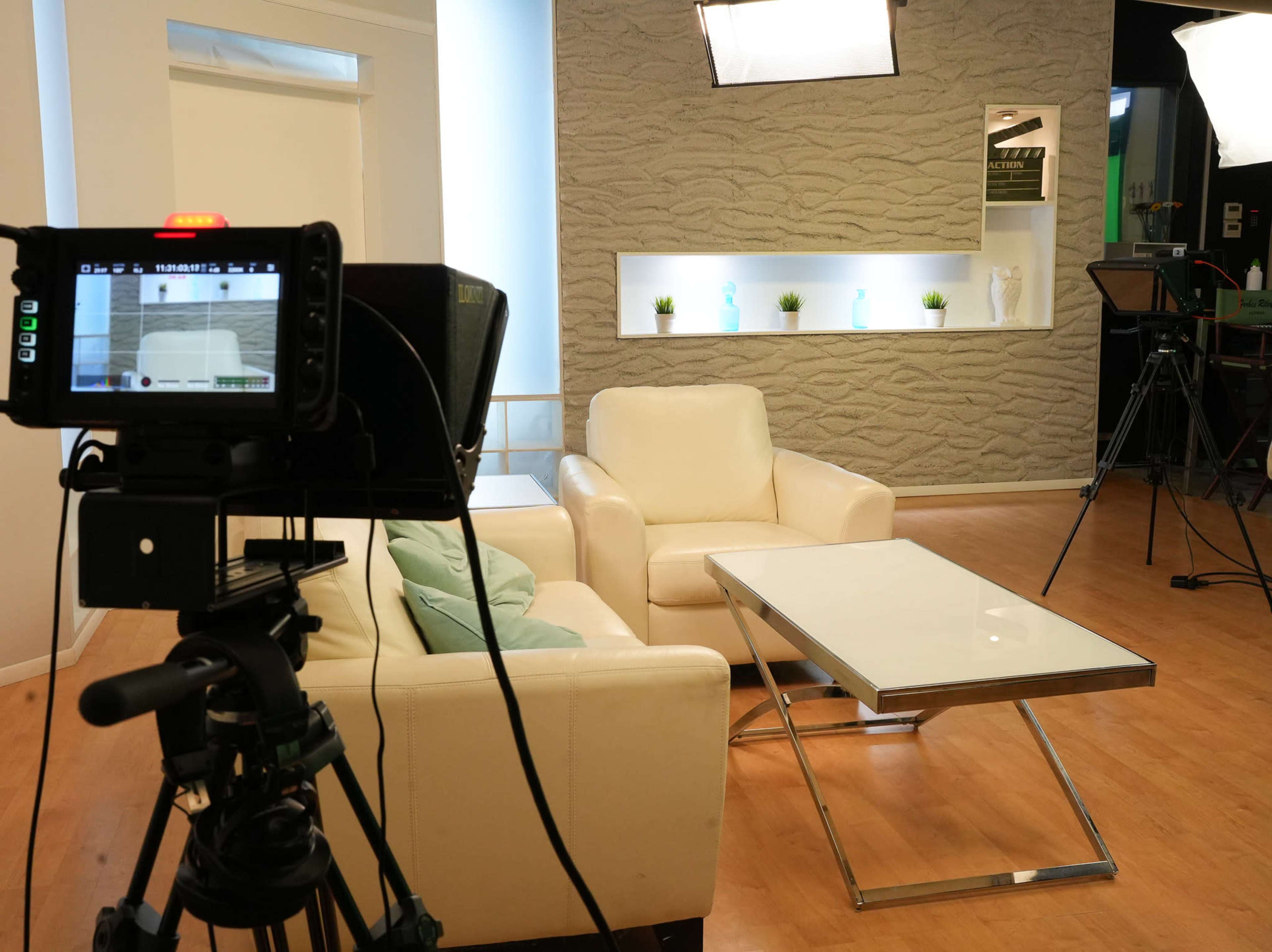 A television studio setup with a camera focused on a white leather couch, a glass coffee table, and soft lighting in the background.