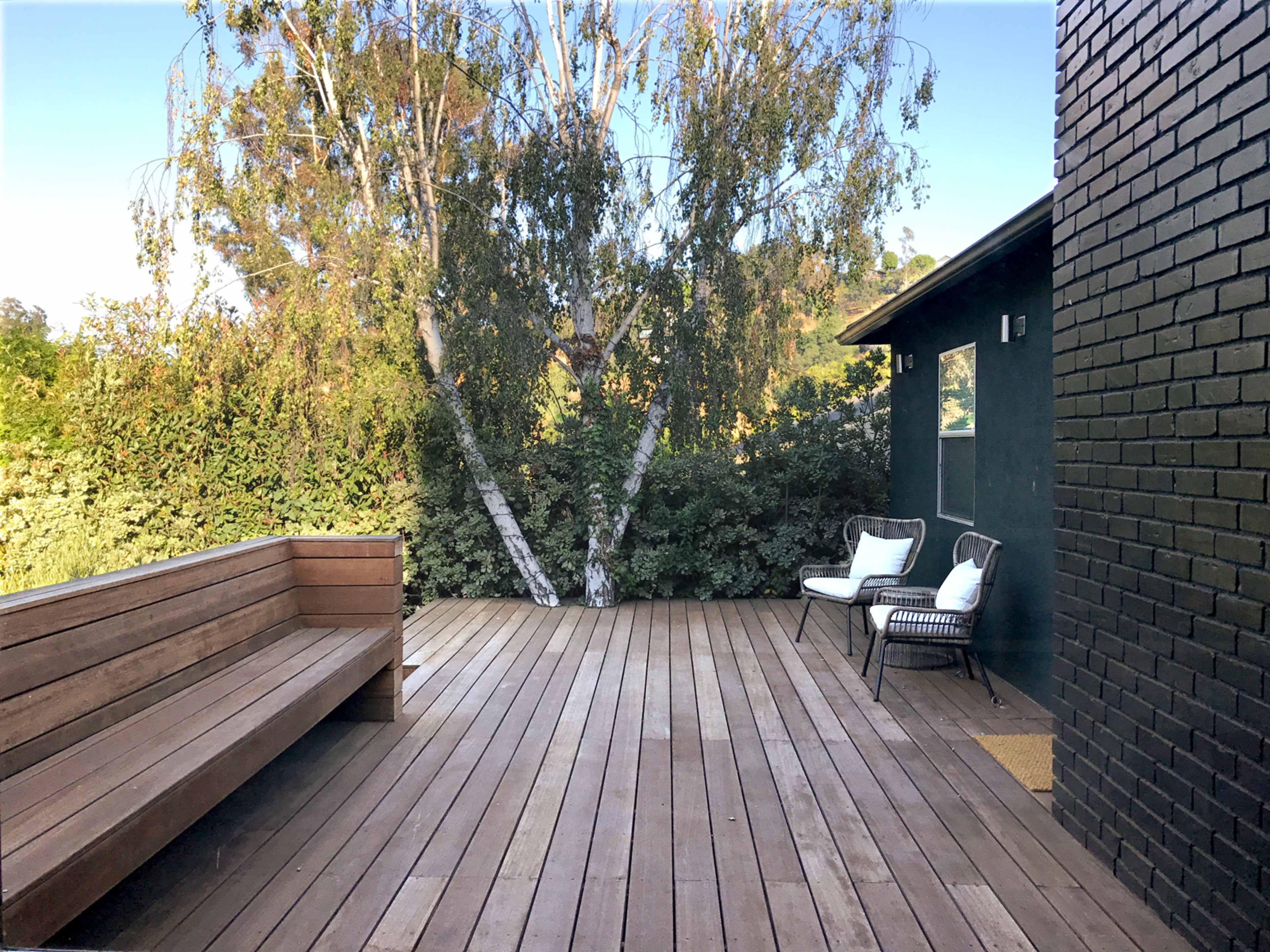 The image shows a wooden deck with a long bench, two chairs, and a backdrop of greenery and a tree.