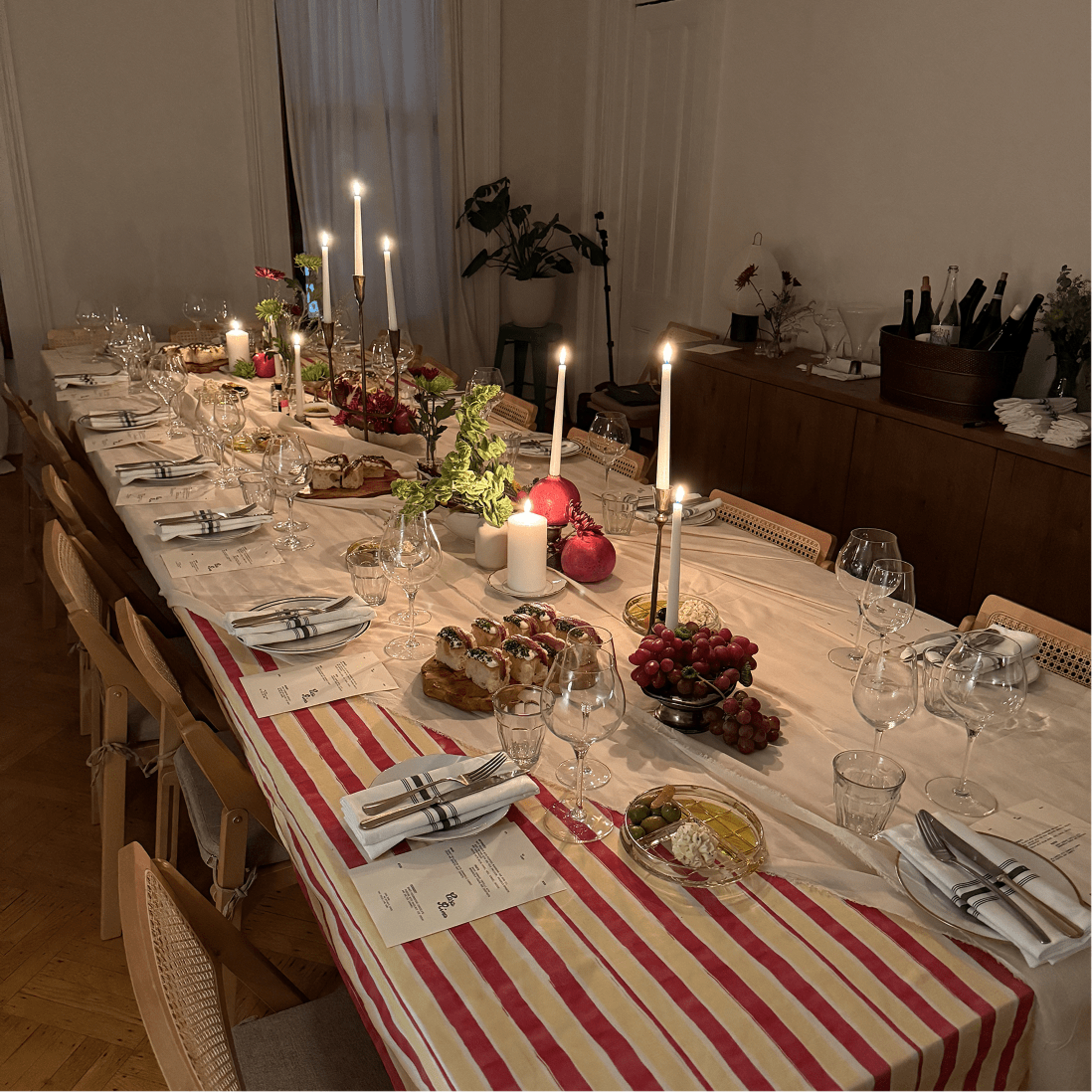 A long dining table is set with elegant glassware and silverware, adorned with candles and an assortment of fruits and breads.