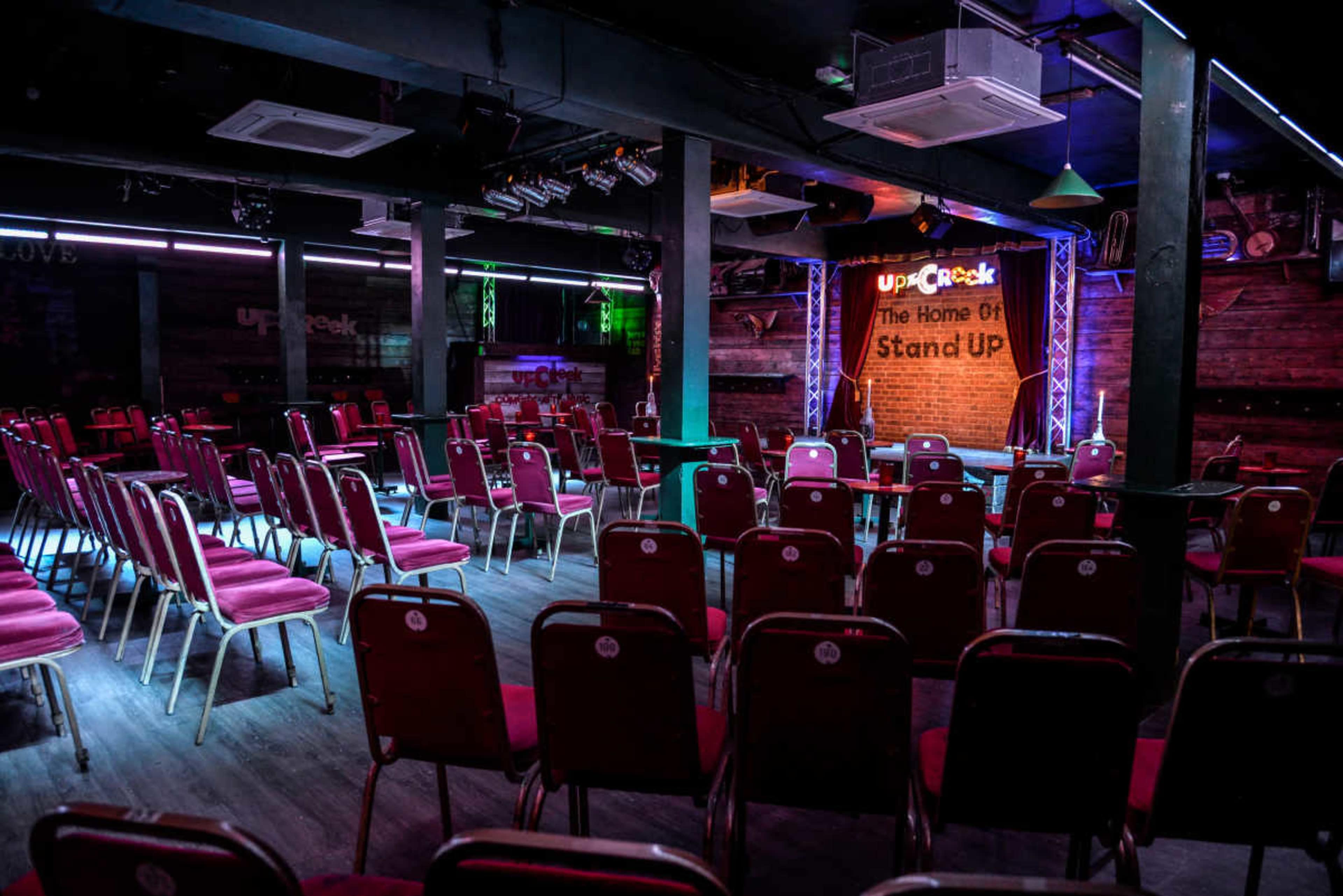 The image shows an empty stand-up comedy venue with rows of red chairs facing a stage illuminated by colorful lights and a sign that reads "The Home Of Stand Up."