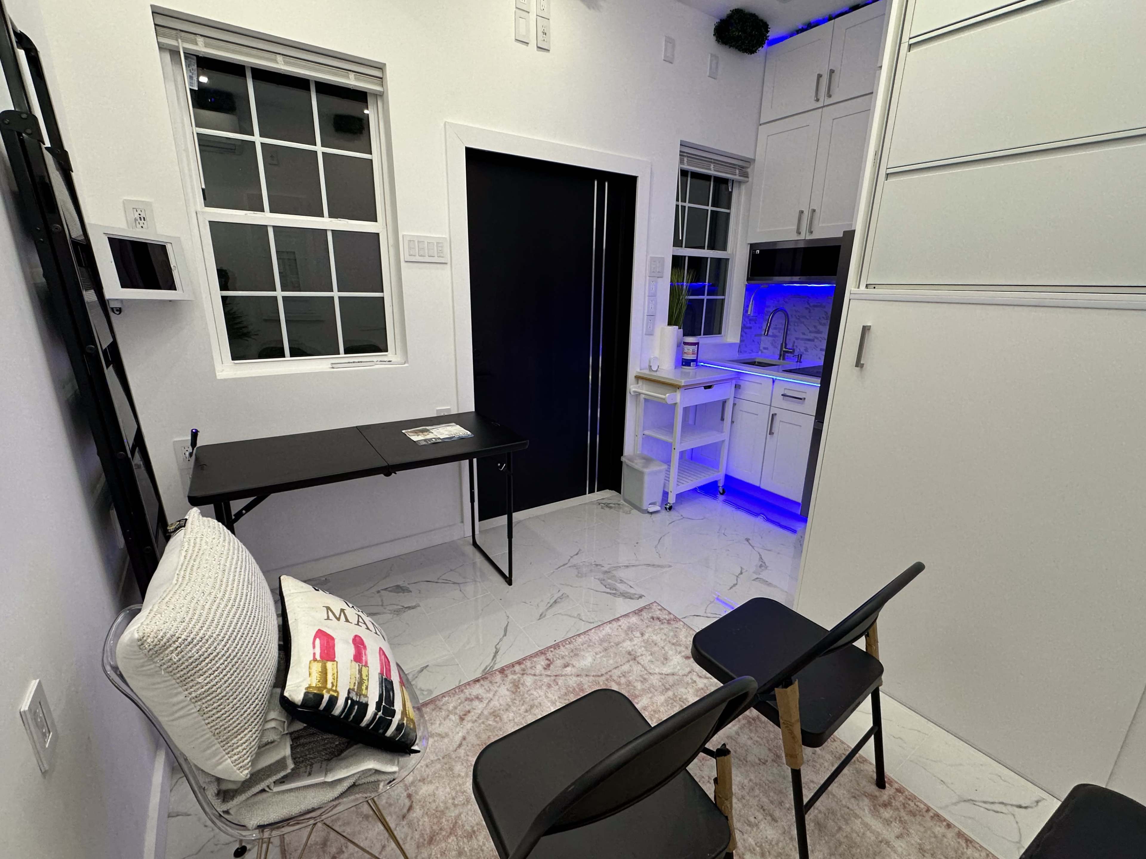 The image shows a small, modern room featuring a kitchenette, a black door, a table, and two chairs on a tiled floor.