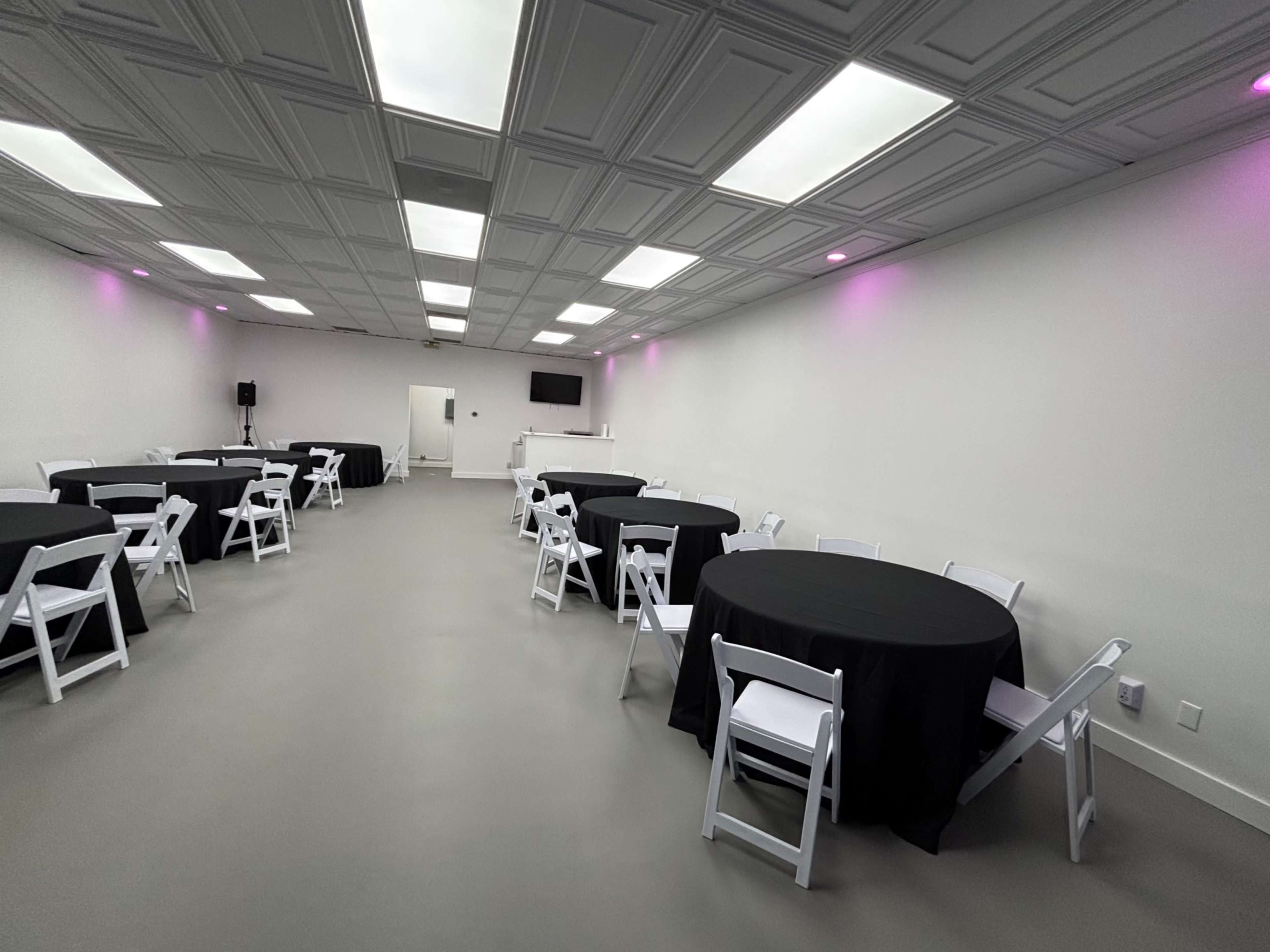 The image shows a spacious, well-lit event room with round tables covered in black cloth and white folding chairs arranged around them.