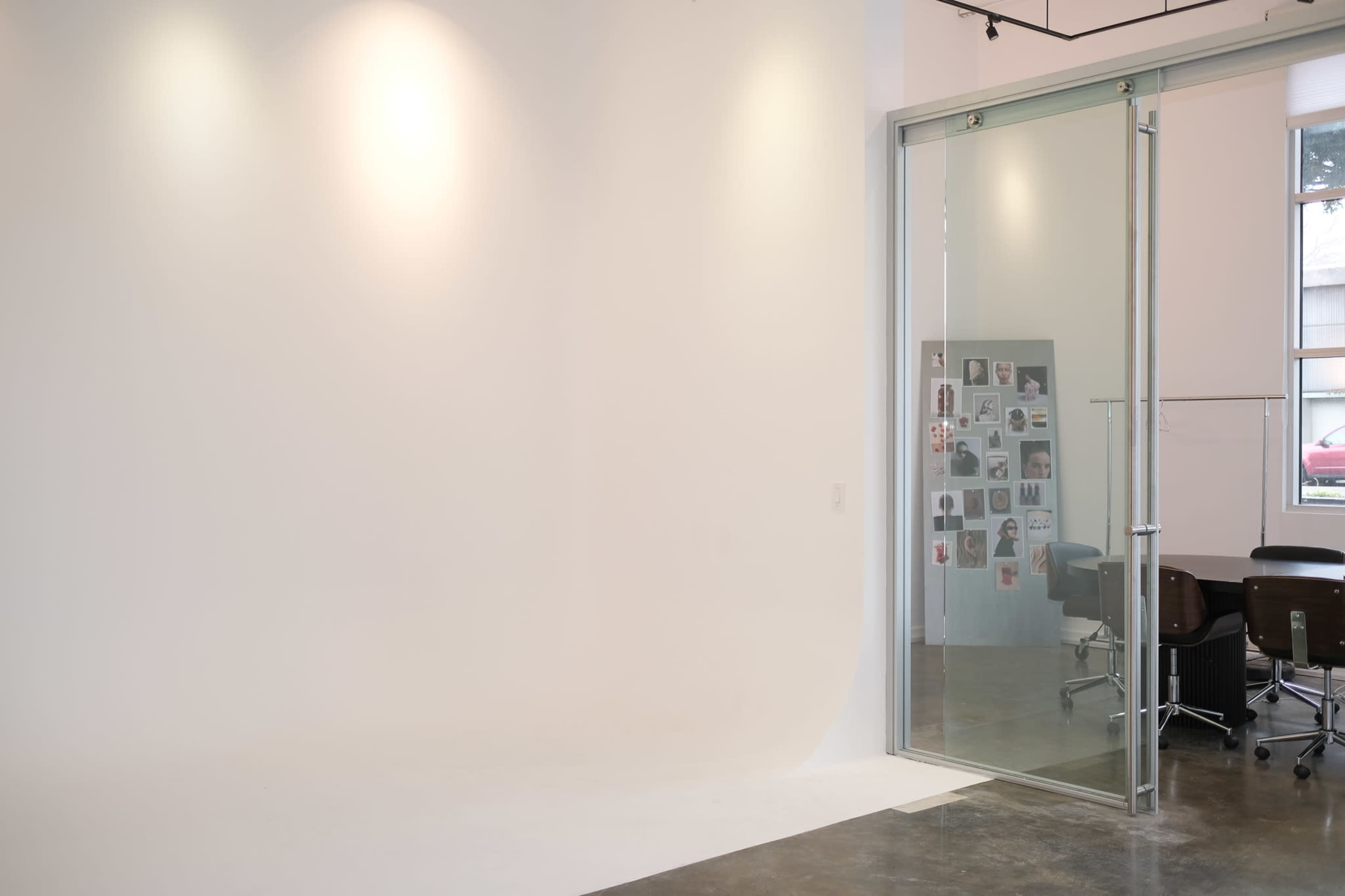 The image shows a minimalist studio space featuring a large white wall, a glass door, and a small meeting area with a table and chairs.