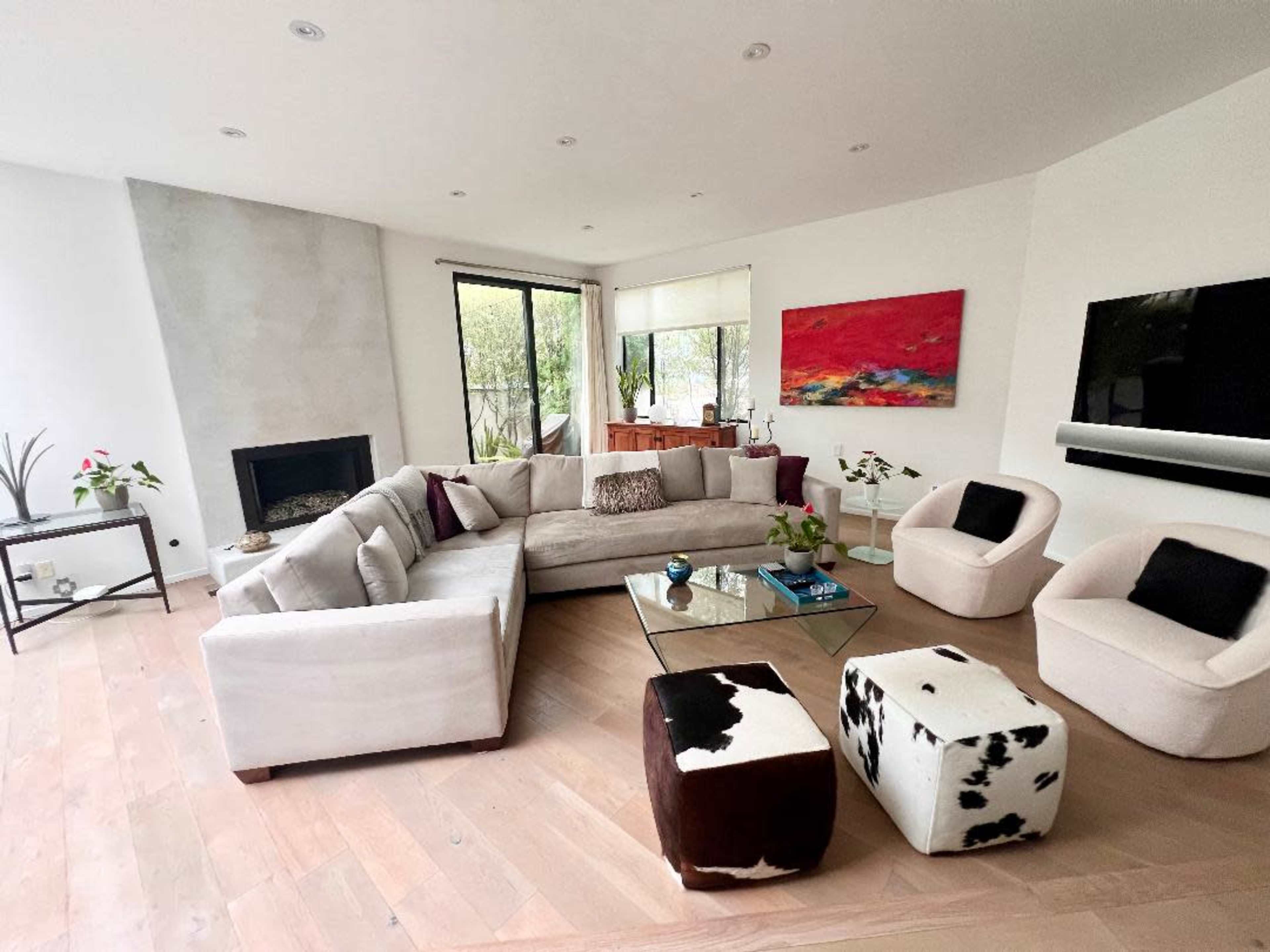 A spacious living room features a large sectional sofa, a glass coffee table, and two unique ottomans, with large windows allowing natural light to enter.