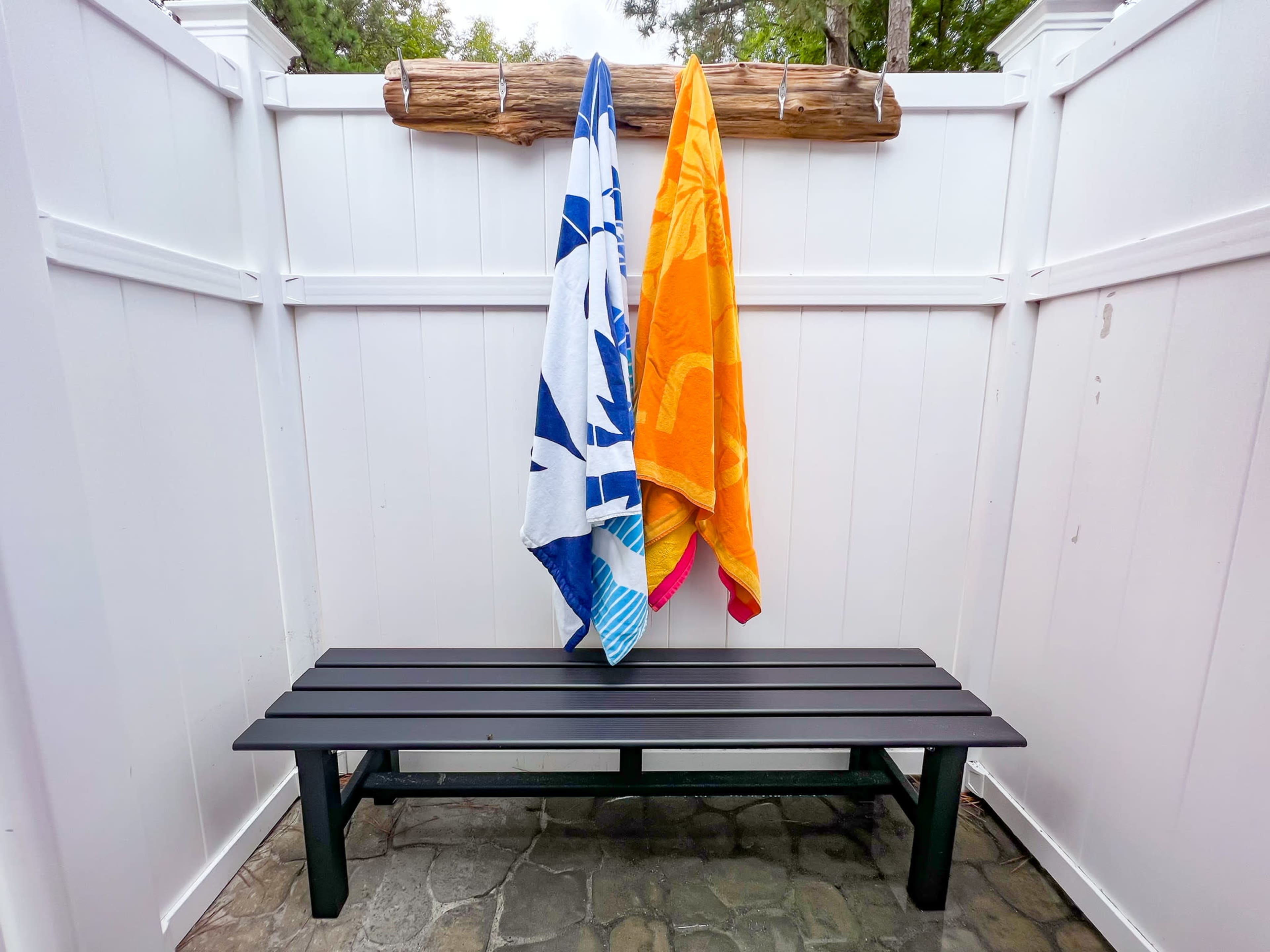Two beach towels hang on a wooden rack above a black bench in an outdoor shower area enclosed by white fencing.