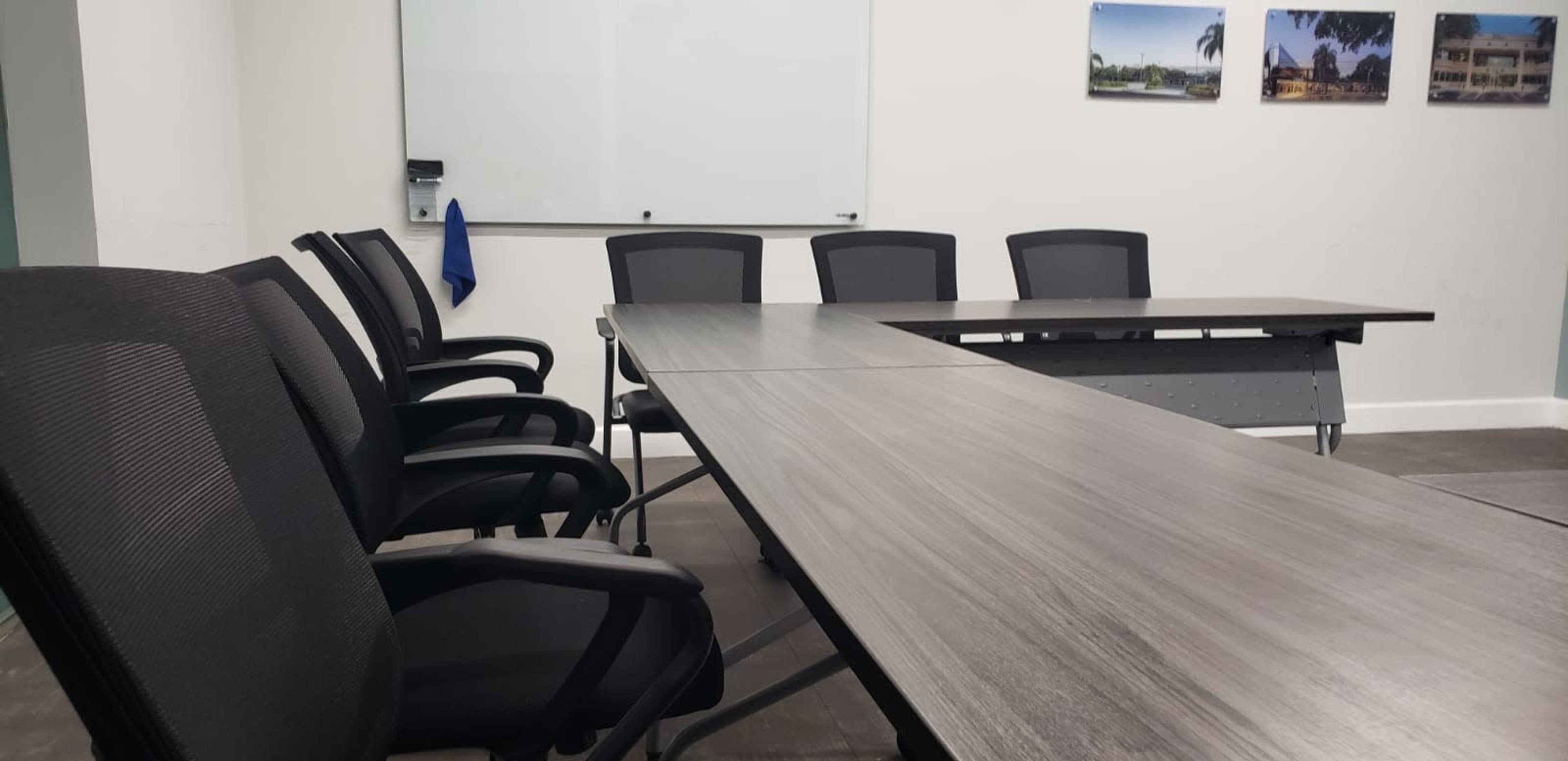 The image shows a conference room featuring a large table surrounded by several black office chairs, with a whiteboard and framed photographs on the wall.