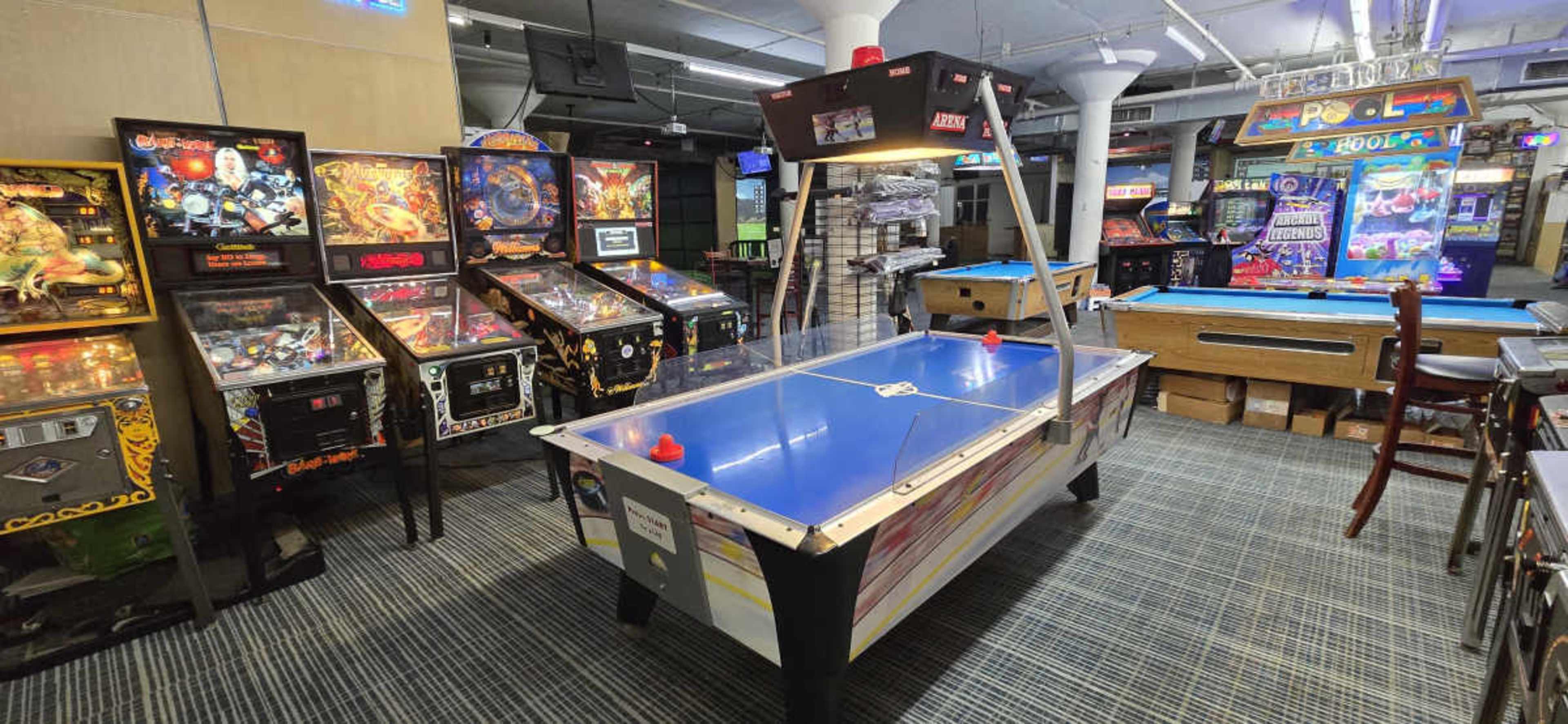 The image shows a game room featuring several pinball machines and an air hockey table in the center, with a pool table visible in the background.