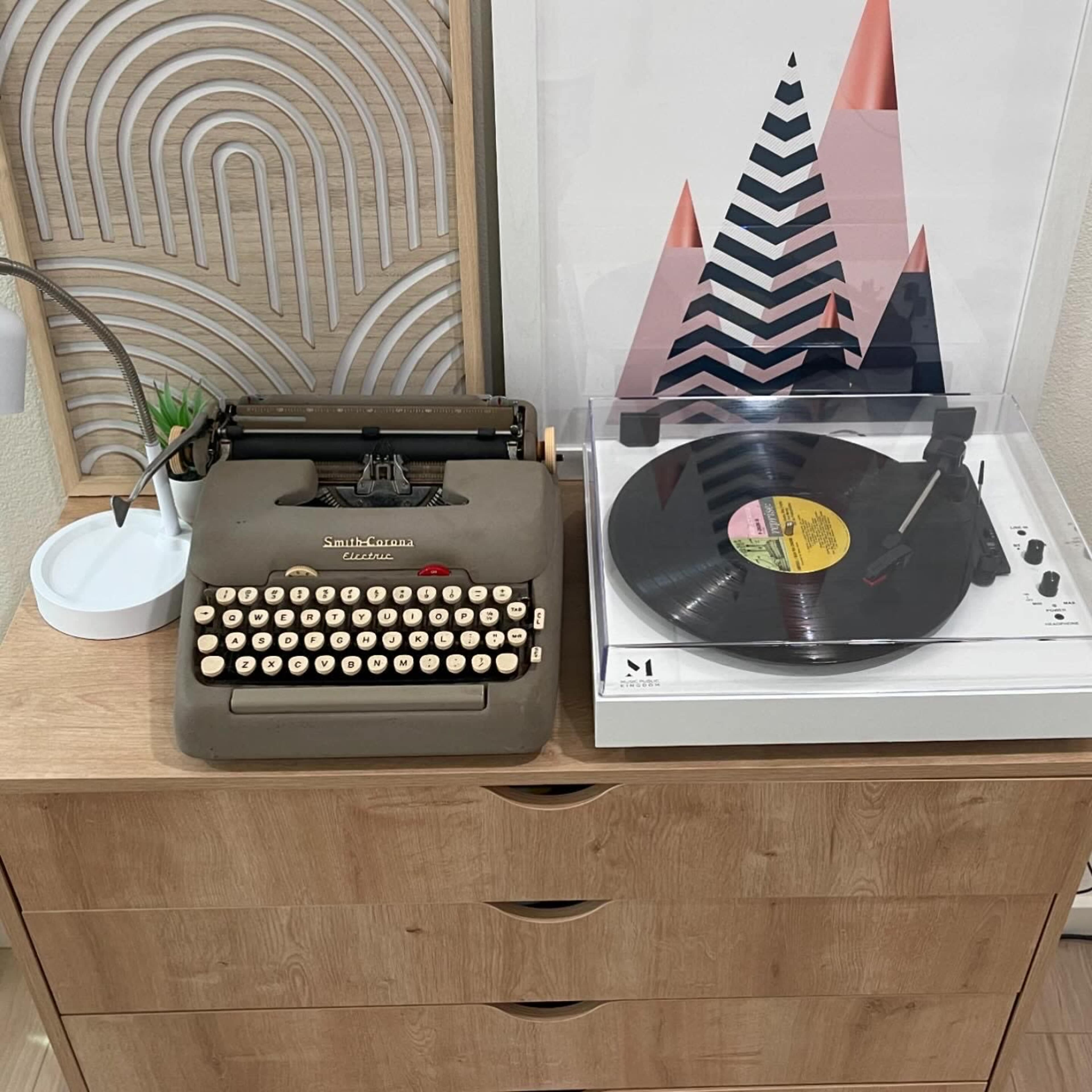 A vintage typewriter sits next to a modern record player on a wooden dresser.