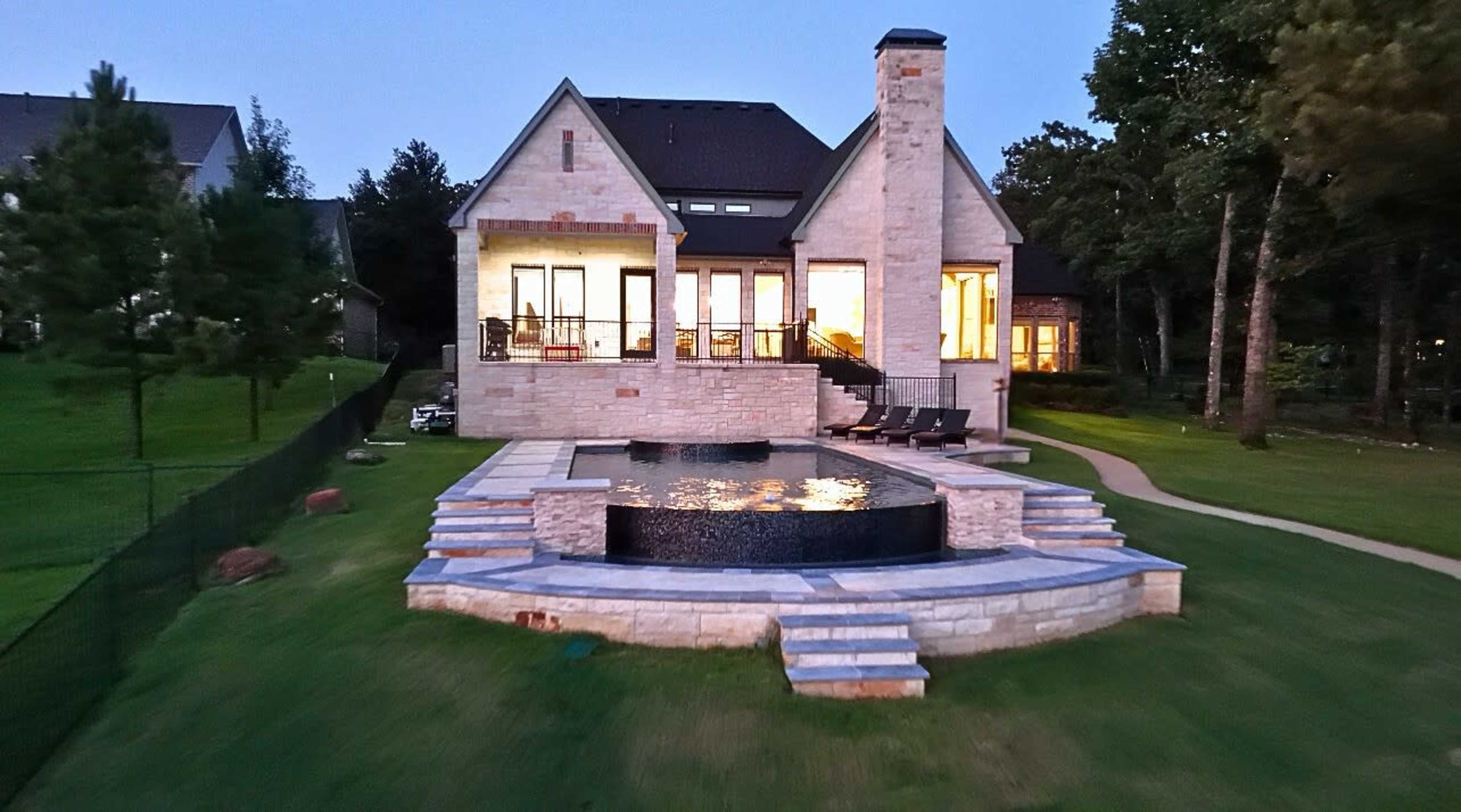 The image shows a modern stone house with a landscaped yard, featuring a circular pool and seating area.