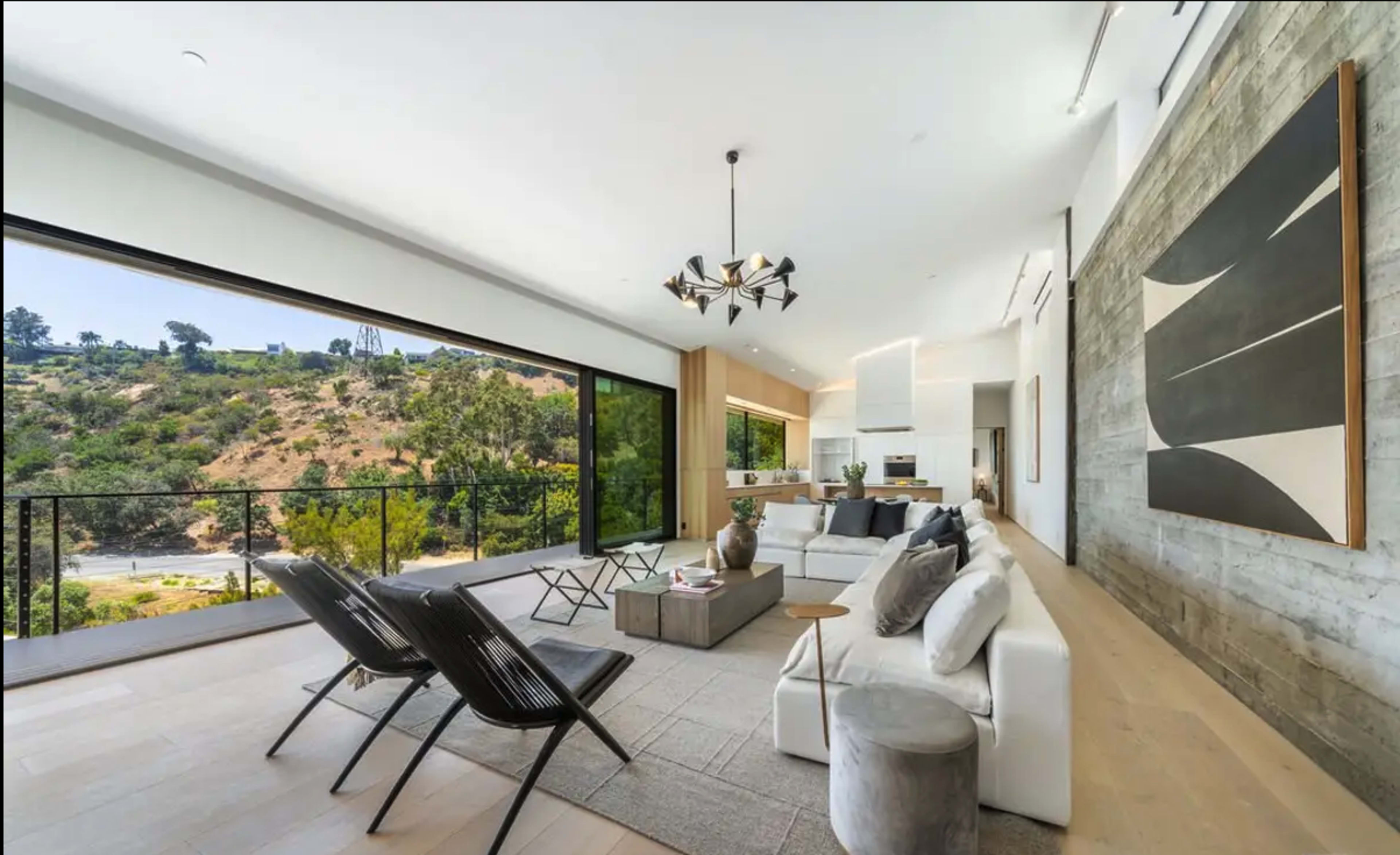 The interior of a modern living room features large windows overlooking a hillside, with a sectional sofa, two lounge chairs, and a minimalist decor.