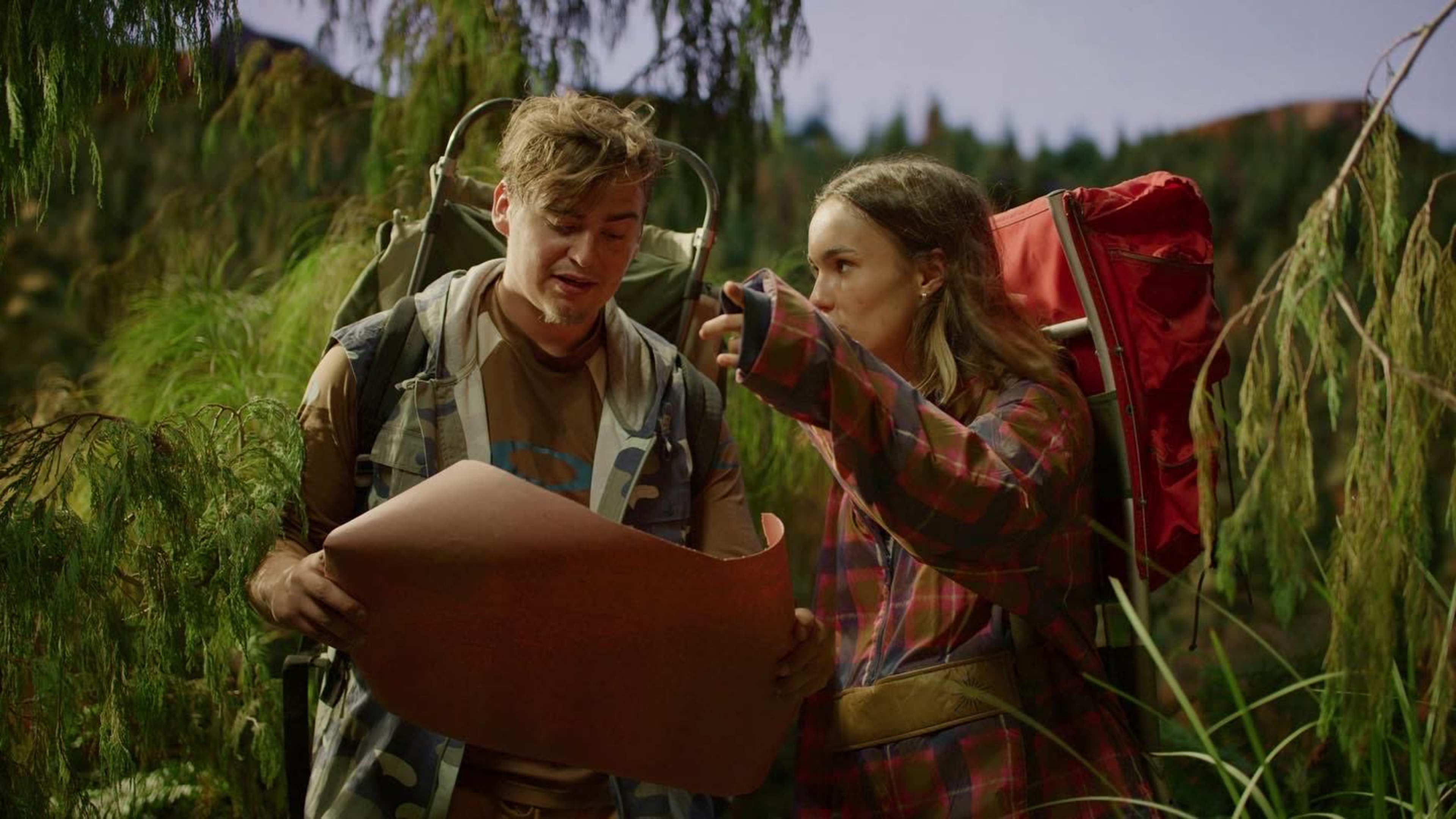 A young man and woman, both wearing backpacks, examine a large map while standing amidst dense greenery.