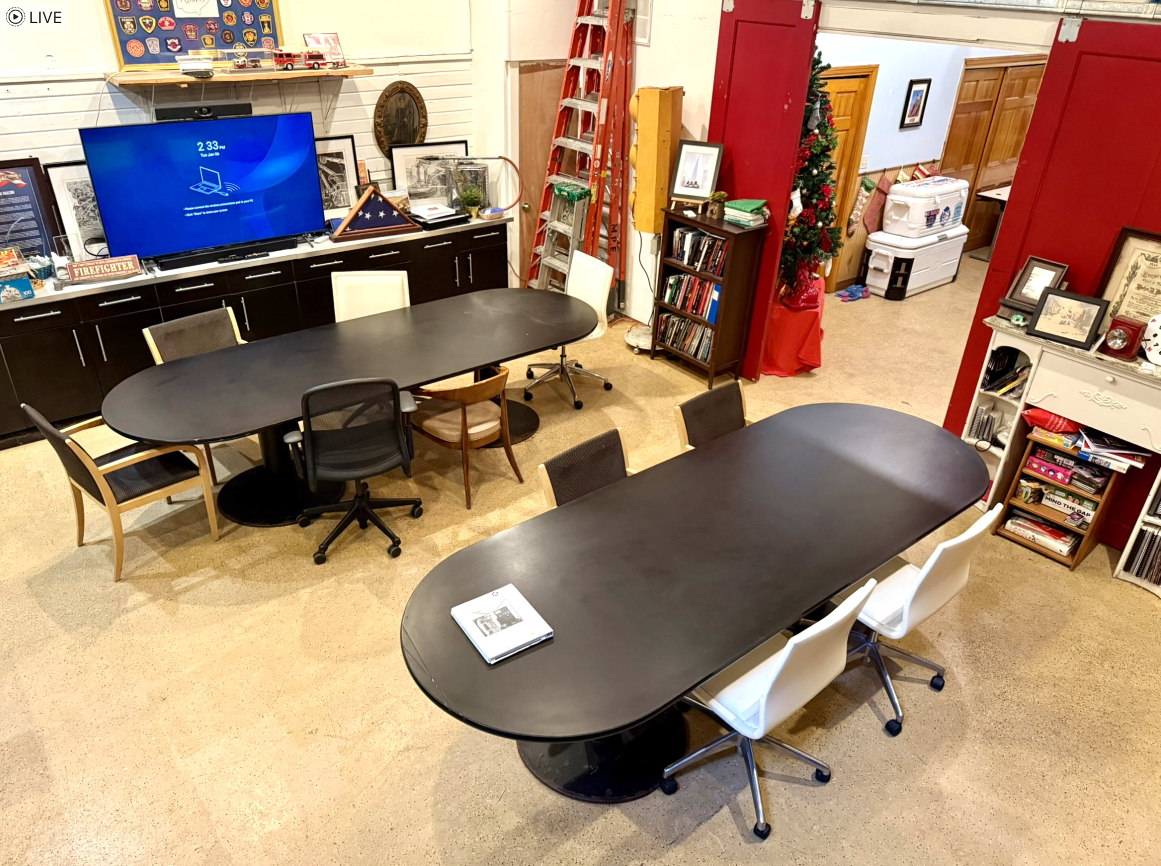 The image shows a spacious room with two large conference tables, a television on the wall, various shelves filled with books and items, and a ladder in the background.