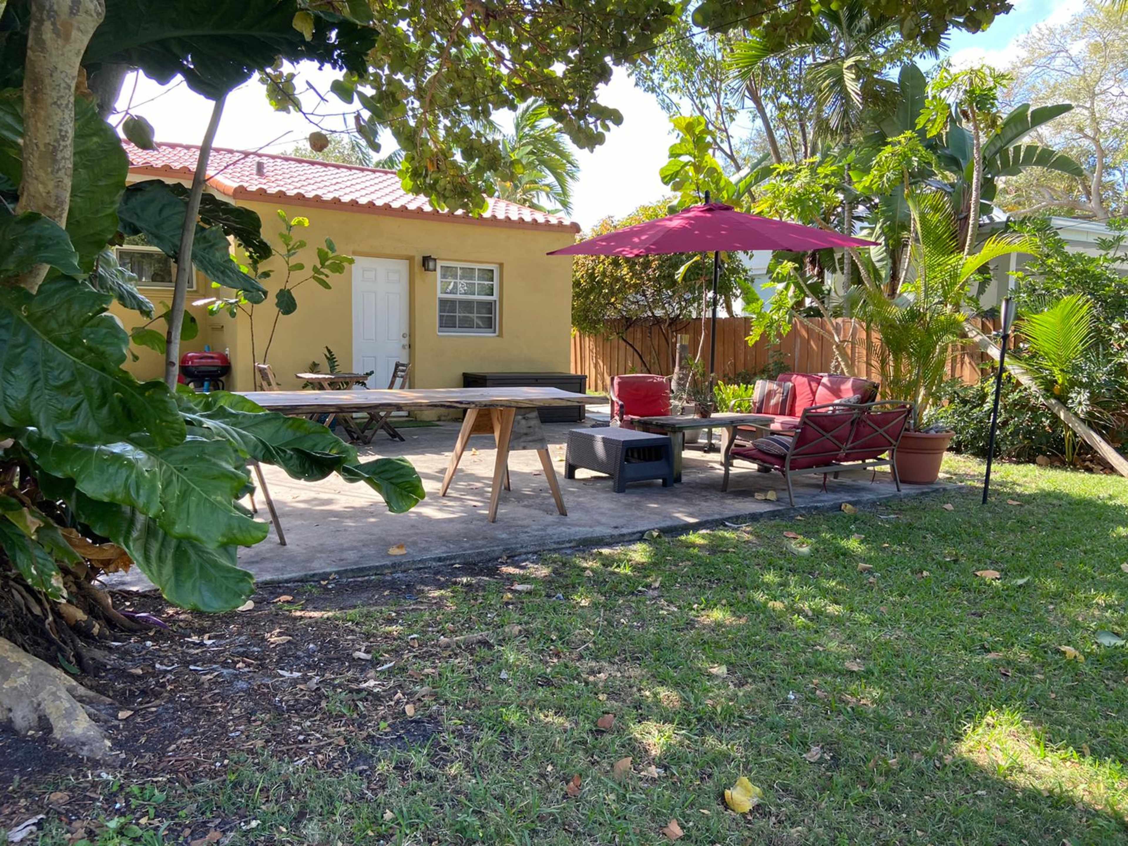 An outdoor patio area with a table, chairs, and an umbrella, adjacent to a house surrounded by greenery.