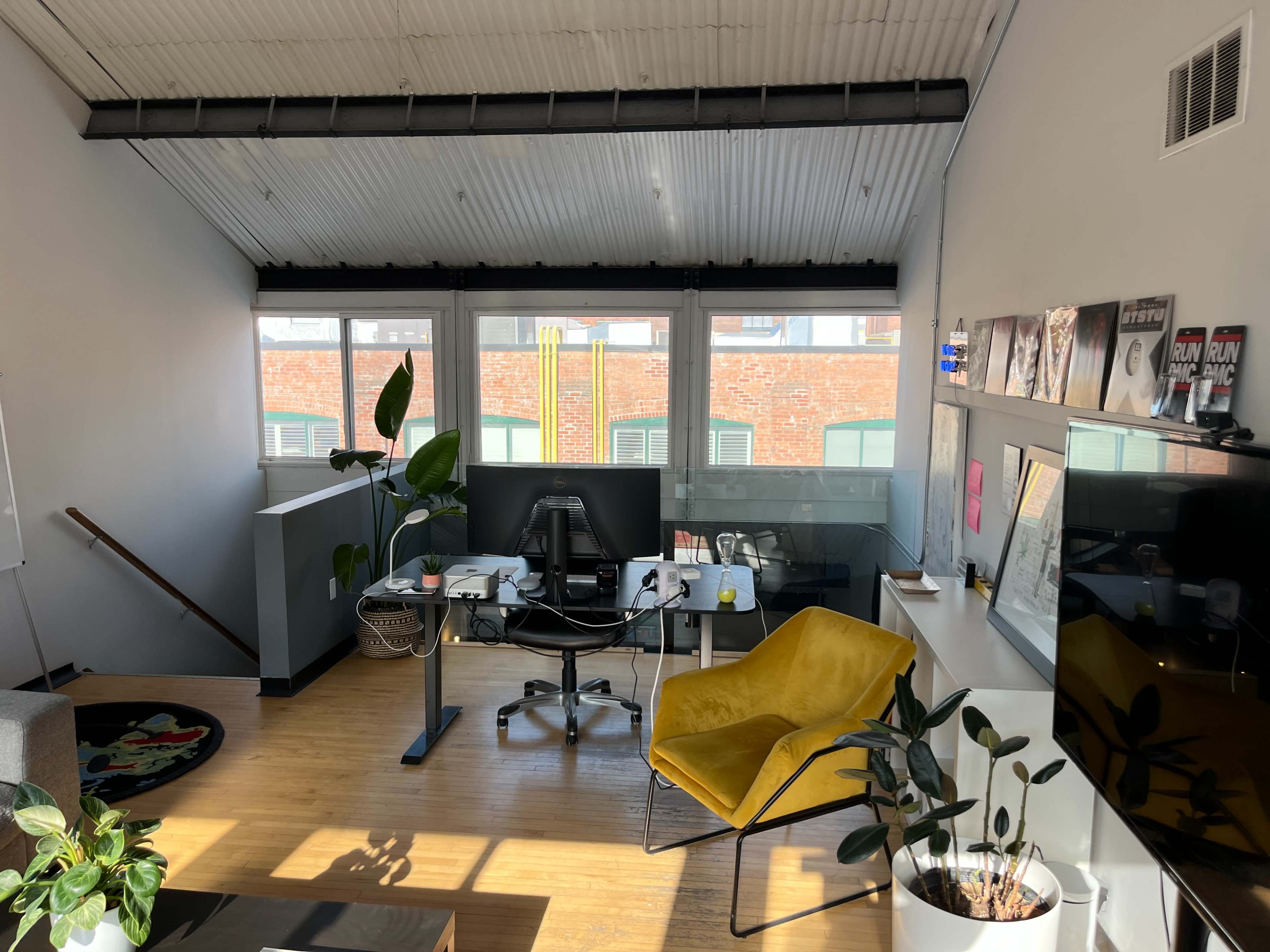 The image shows a modern office space with a desk, computer, yellow chair, plants, and large windows along one wall.