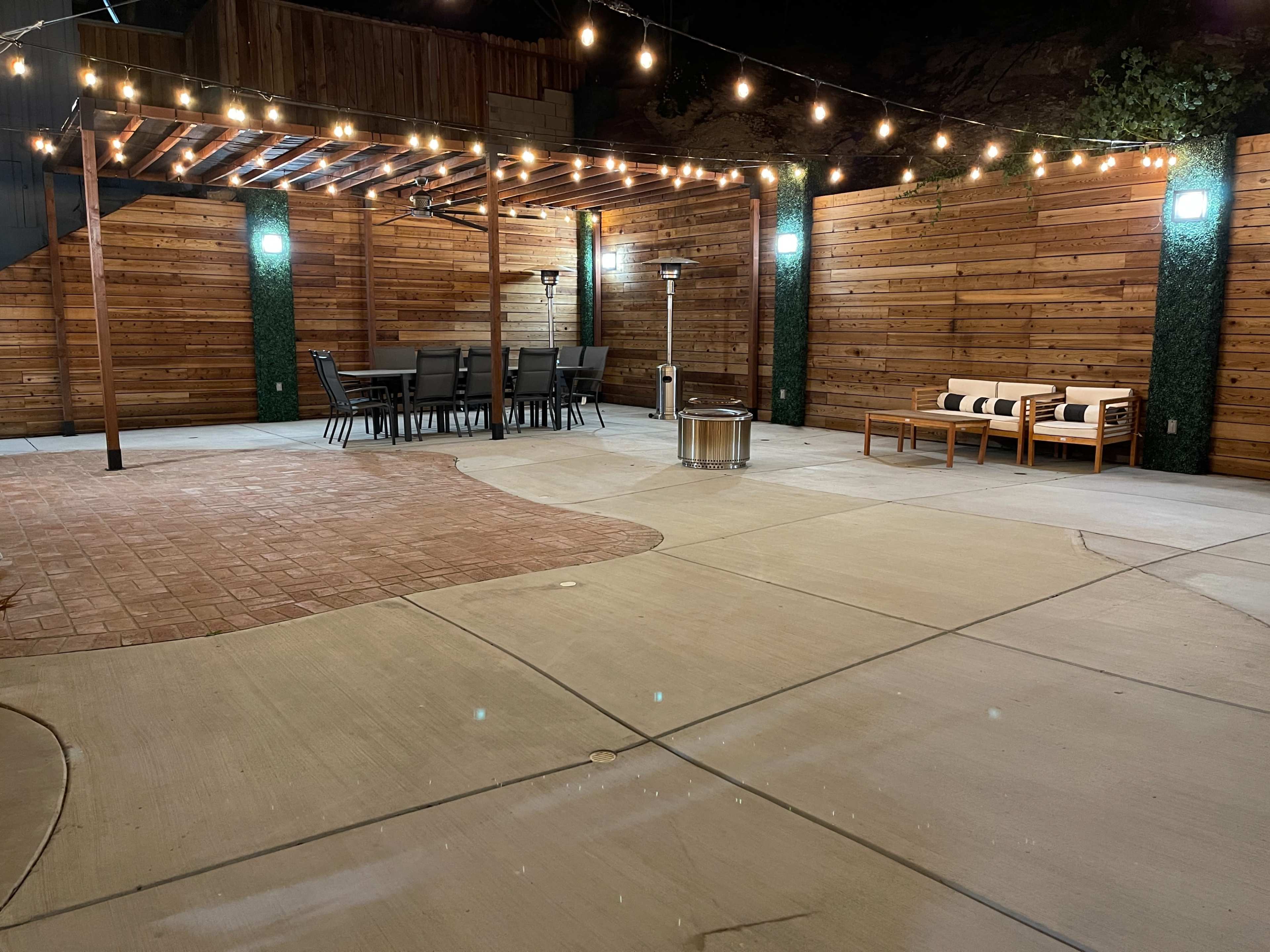 A spacious outdoor patio area at night, featuring a seating arrangement under a pergola with string lights, a dining table, and a fire pit.