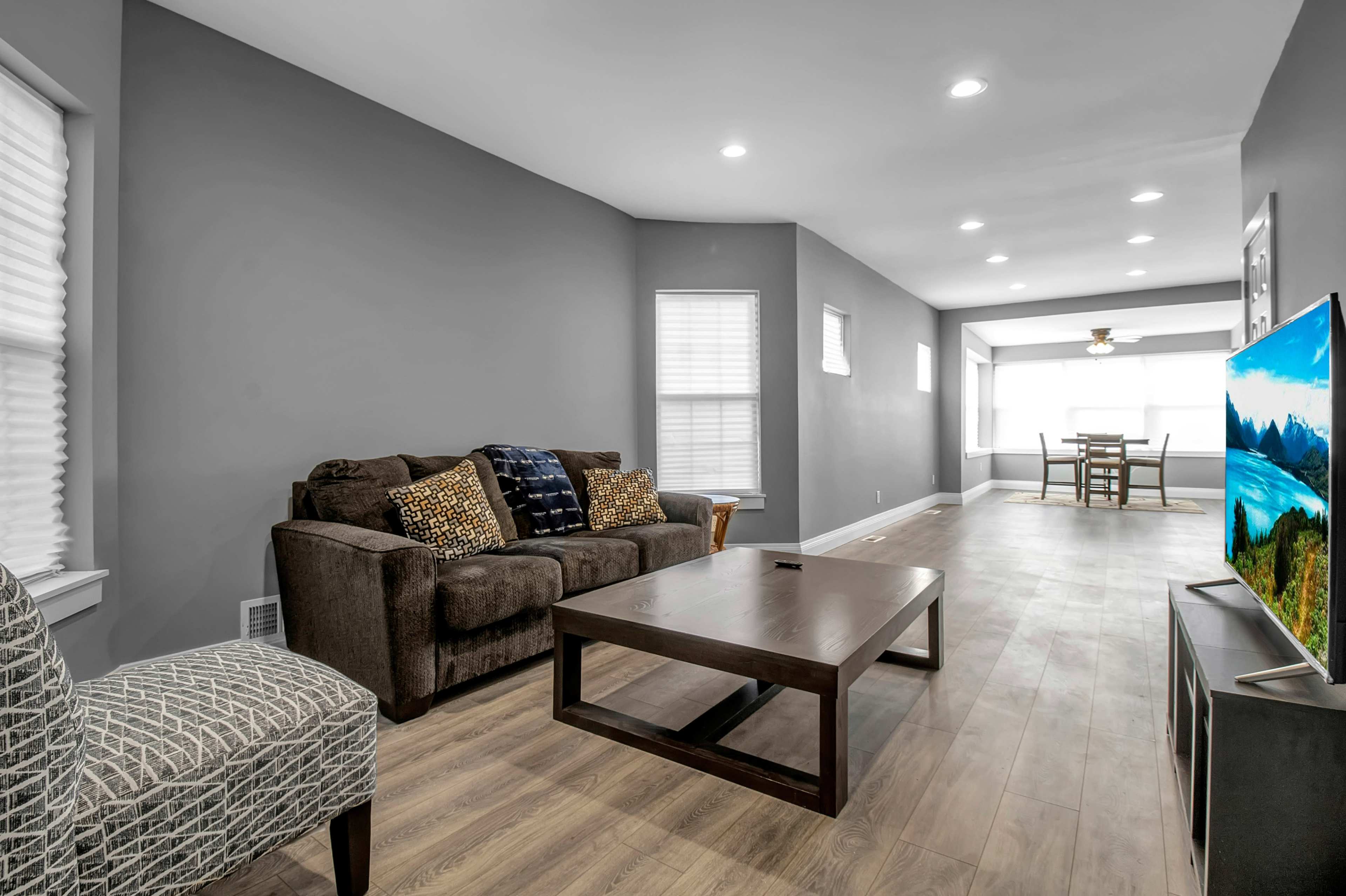 A living room features a brown couch with decorative pillows, a wooden coffee table, and a TV across from a dining area with a ceiling fan and large windows.