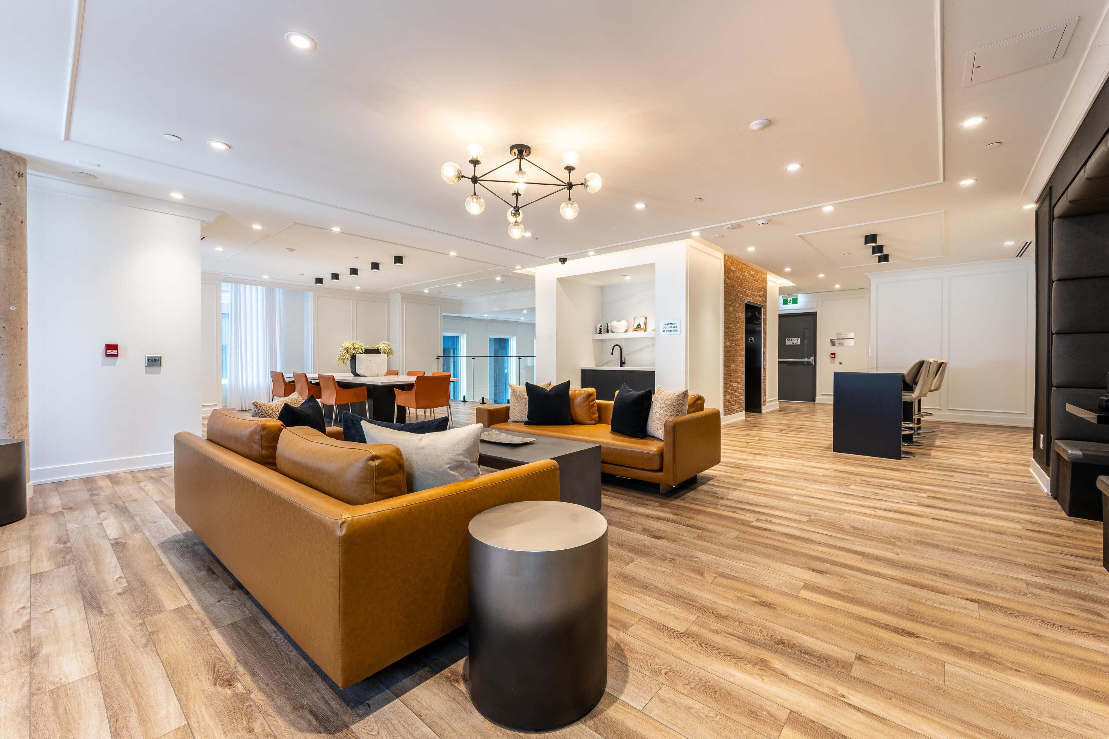 The image shows a modern open-concept living space featuring a brown leather sofa, a round side table, and a dining area with a large table and chairs, all complemented by wood flooring and stylish light fixtures.