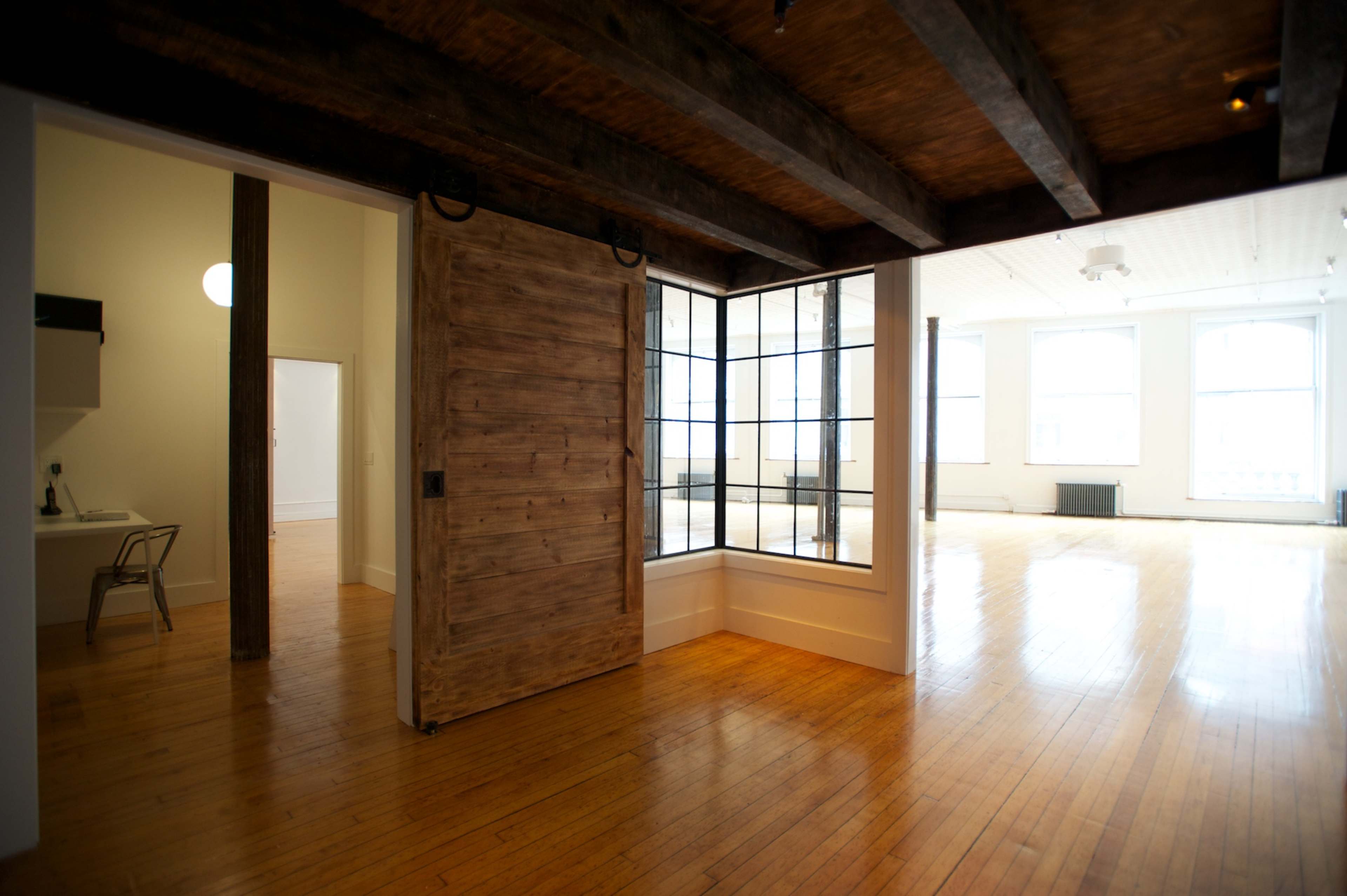 A spacious, well-lit room with wooden floors, large windows, and a sliding wooden door that separates it from another area.