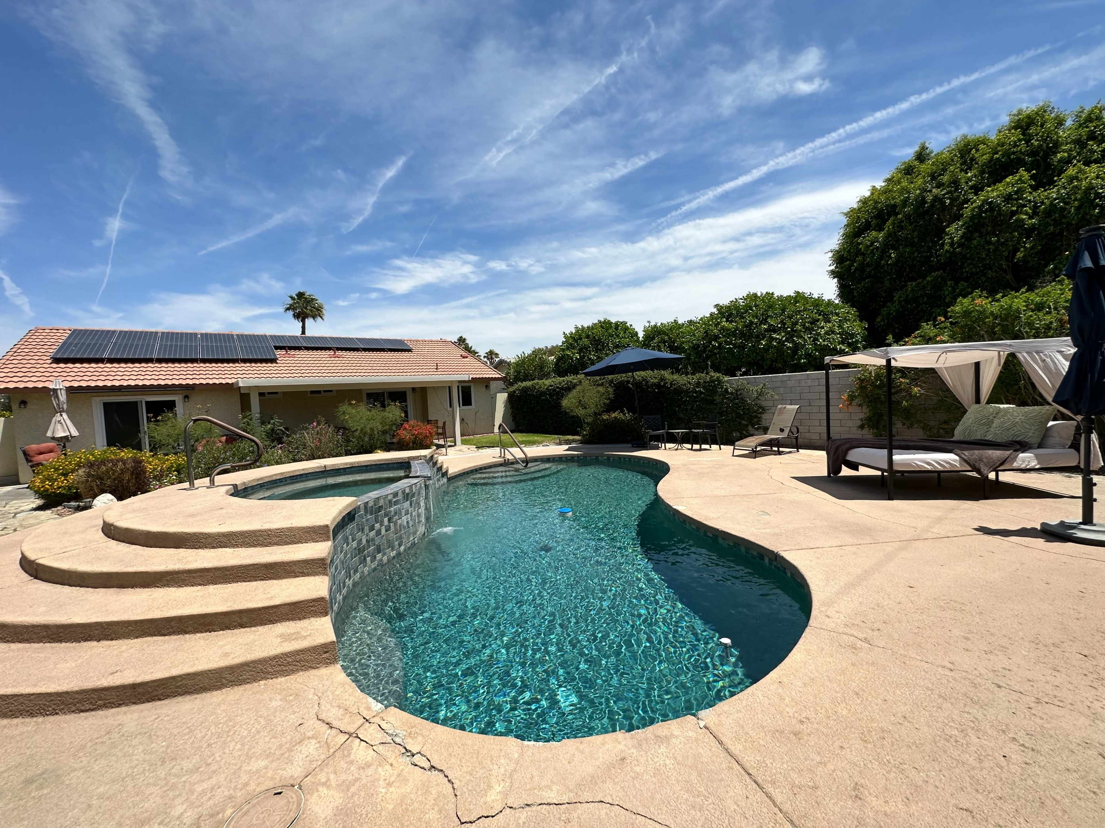 The image features a swimming pool with a spa, surrounded by a patio area with lounge chairs and an umbrella, and a house with solar panels in the background.