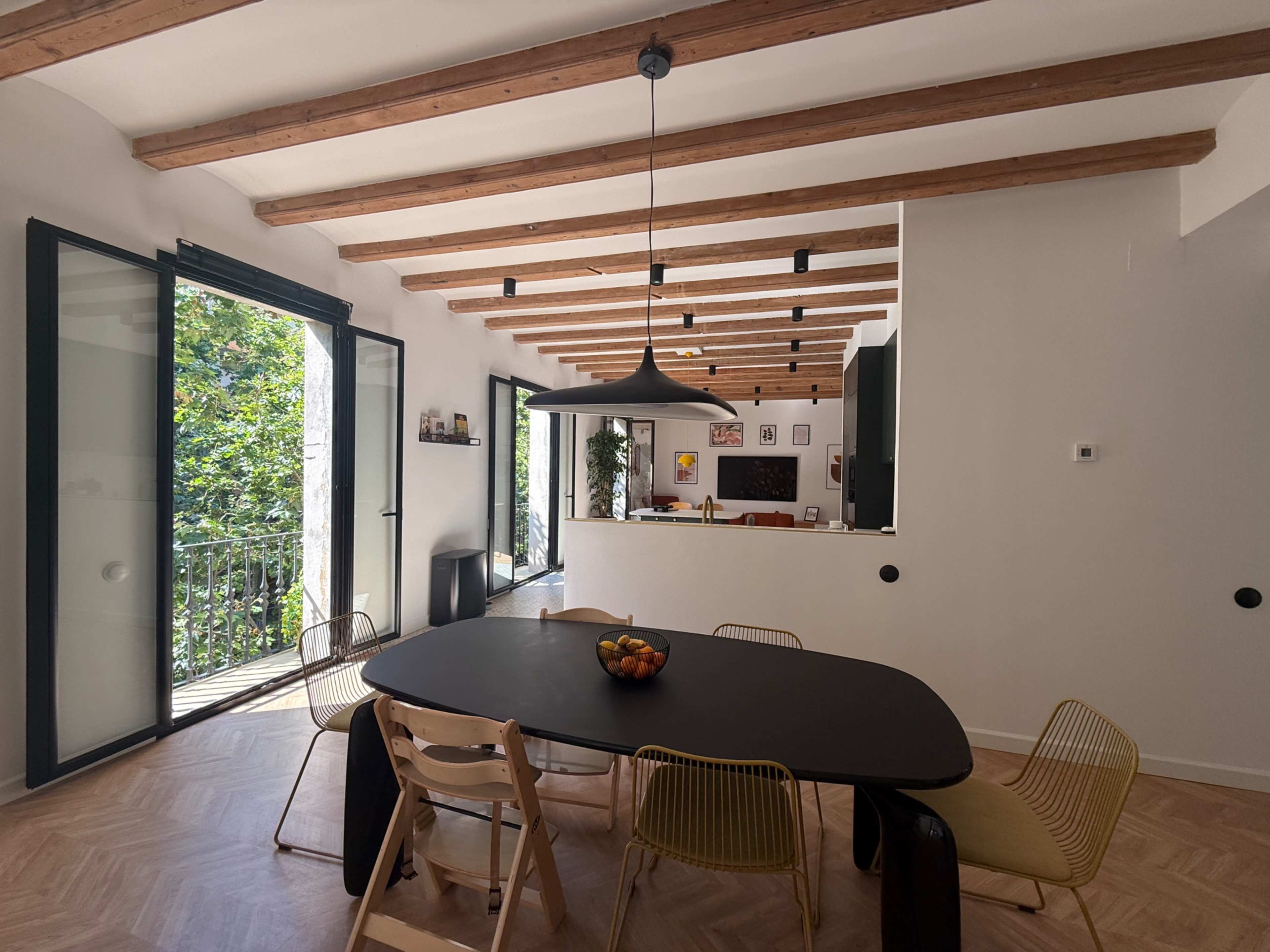 A modern dining area features a round black table surrounded by colorful chairs, with large open doors leading to a view of greenery outside.