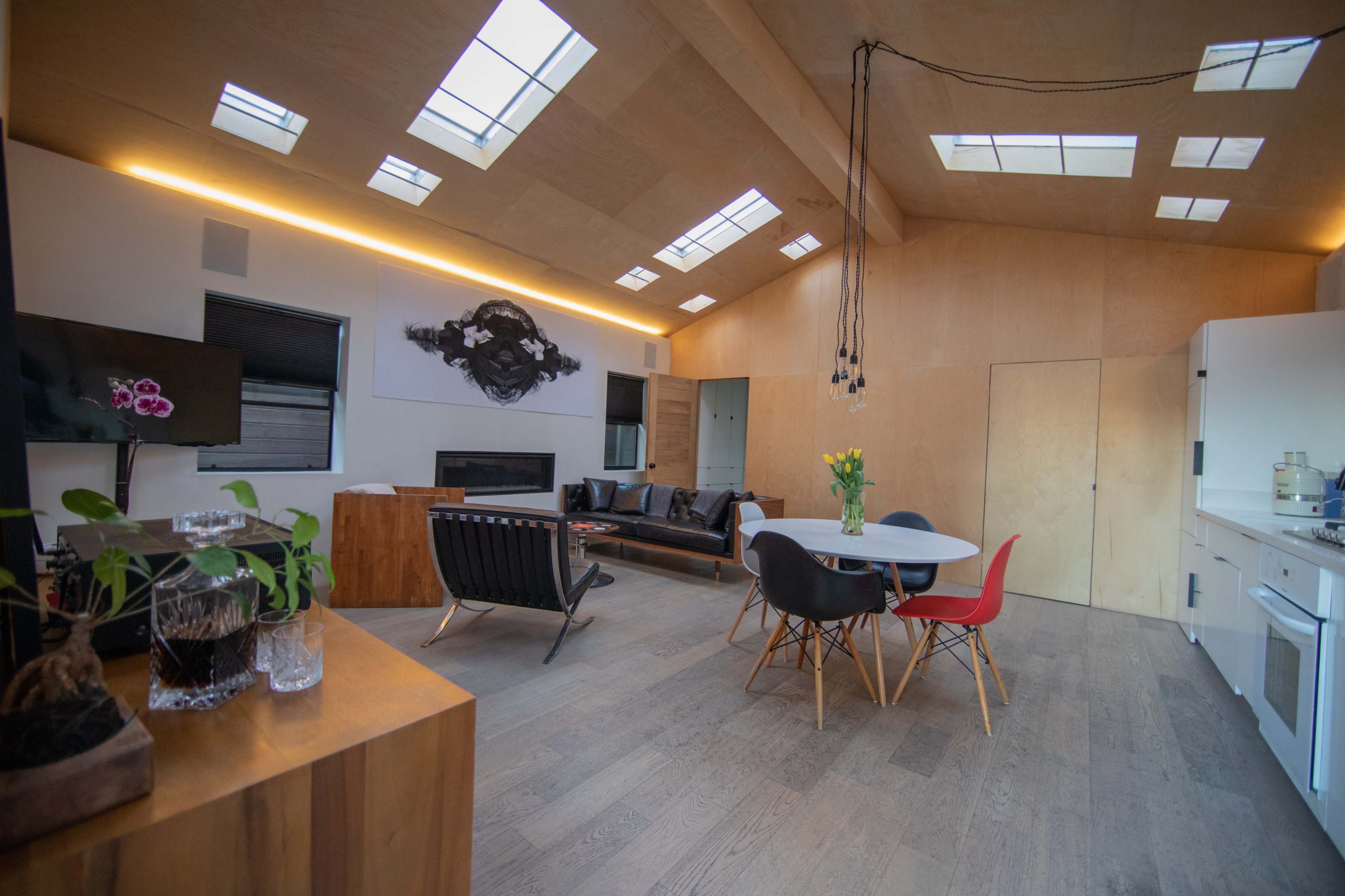 The image shows a modern interior space featuring a living area with a black sofa, a dining table surrounded by colorful chairs, and large windows allowing natural light to enter.