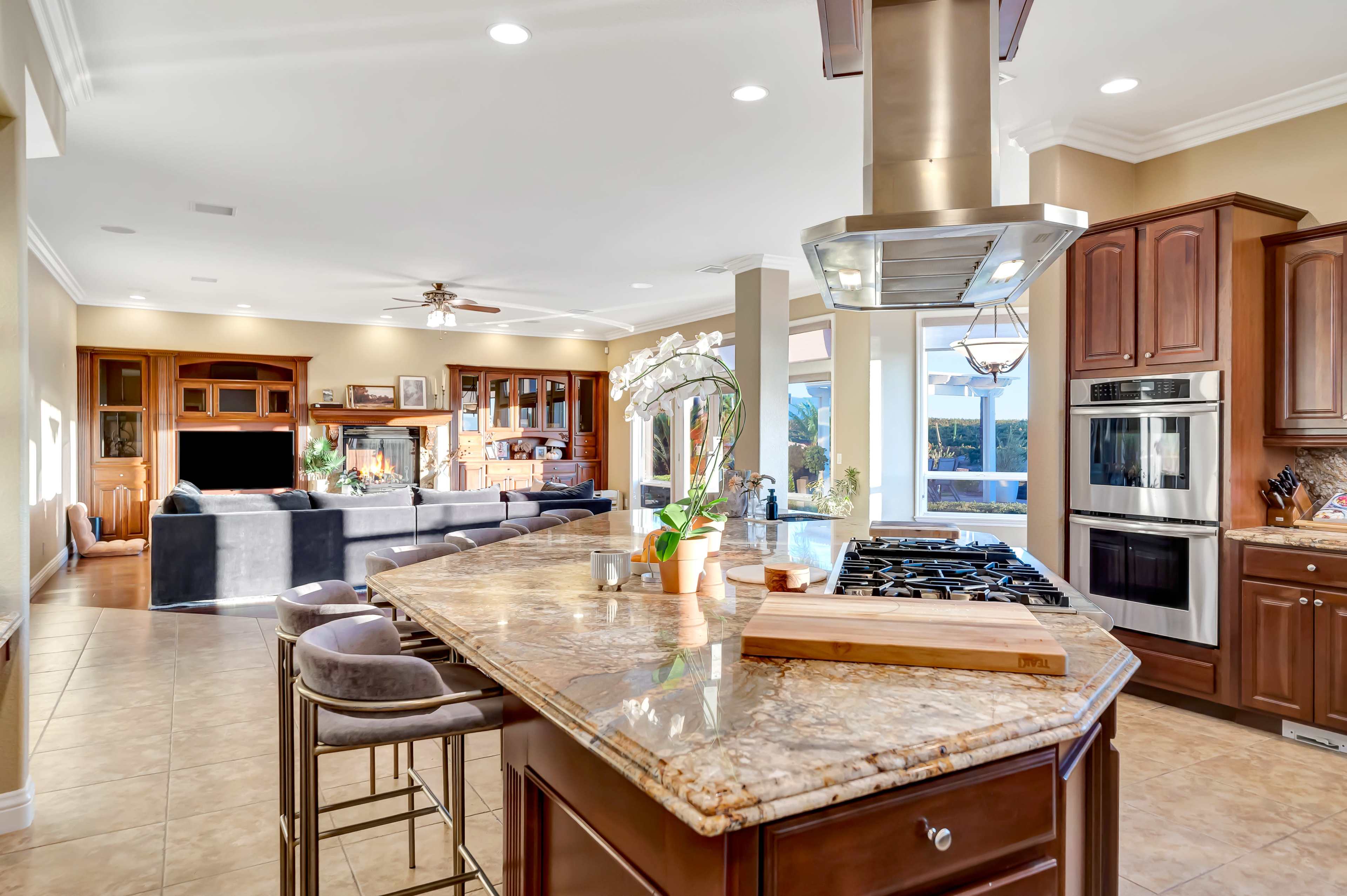 A modern kitchen features a large island with a gas stovetop, surrounded by bar stools, seamlessly connected to a spacious living area with large windows and hardwood cabinetry.