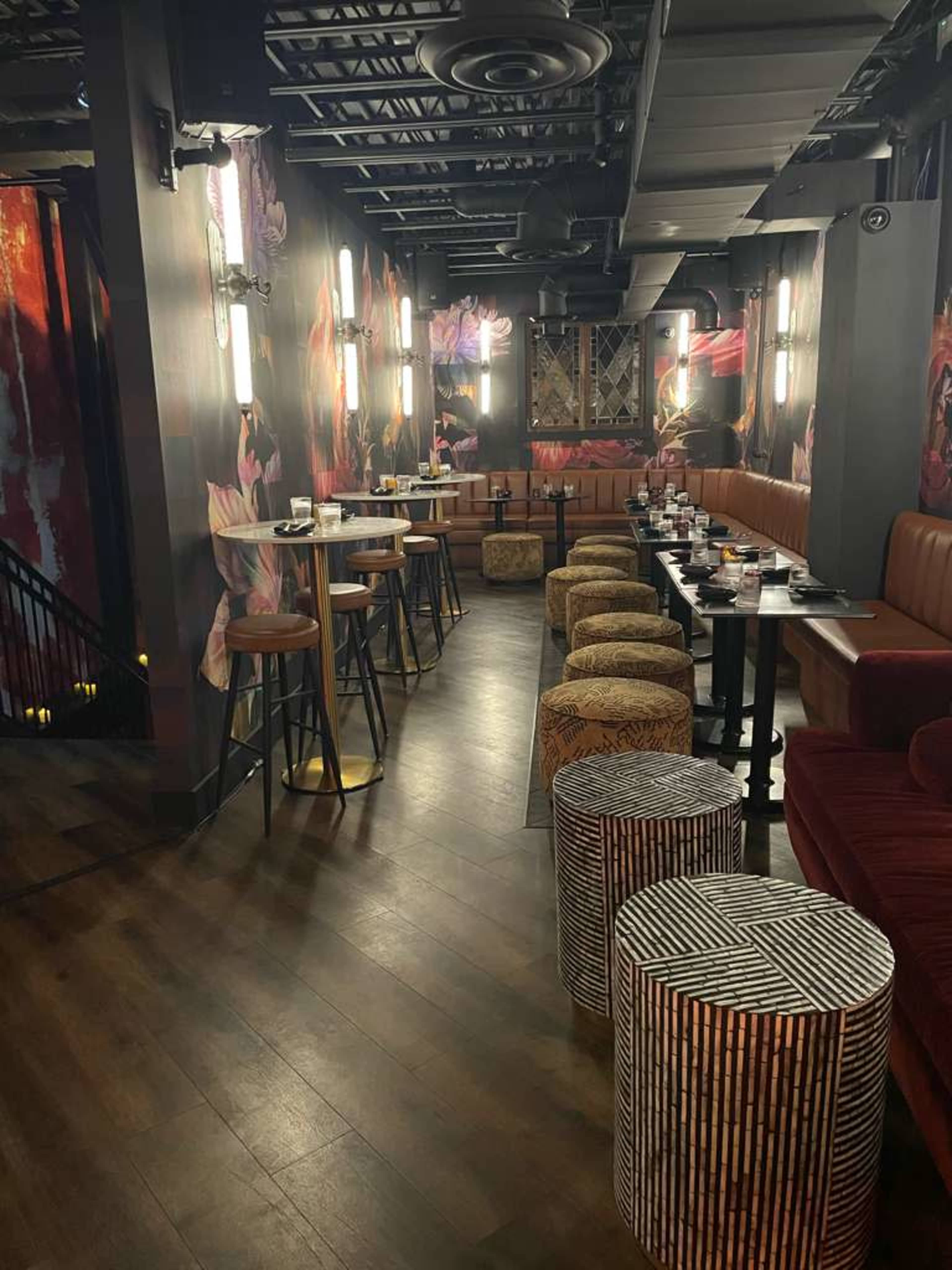 The image shows a stylish bar area with colorful floral wallpaper, wooden flooring, and a mix of high tables and patterned stools.