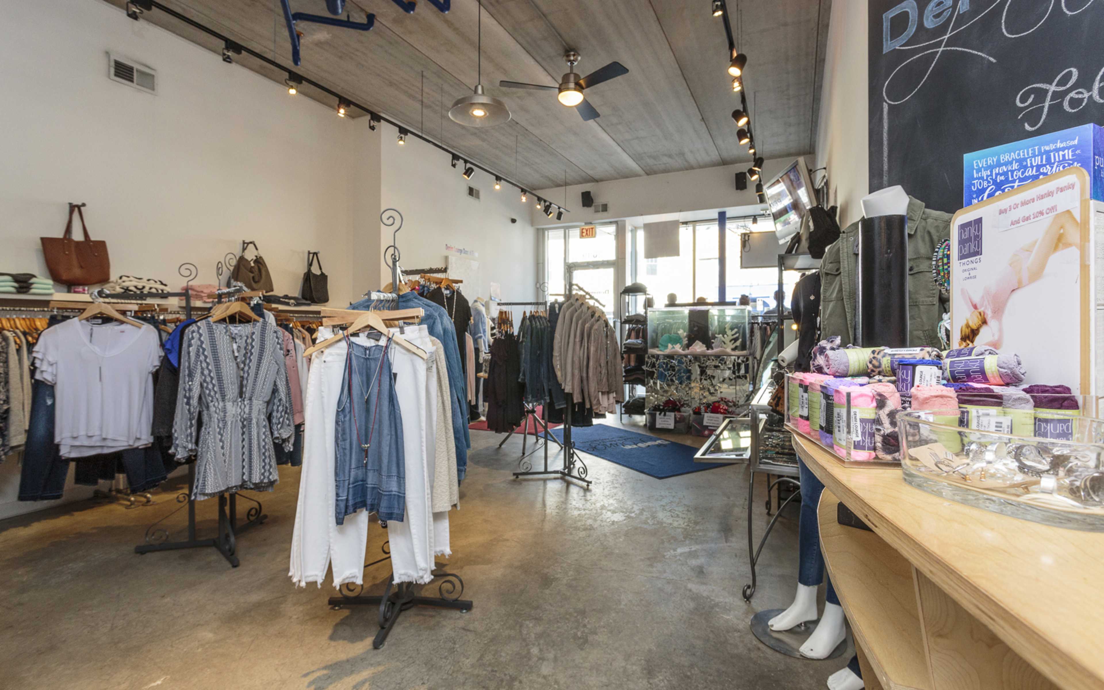 The interior of a clothing store features racks of apparel, mirrors, and a display table with yarn and accessories.