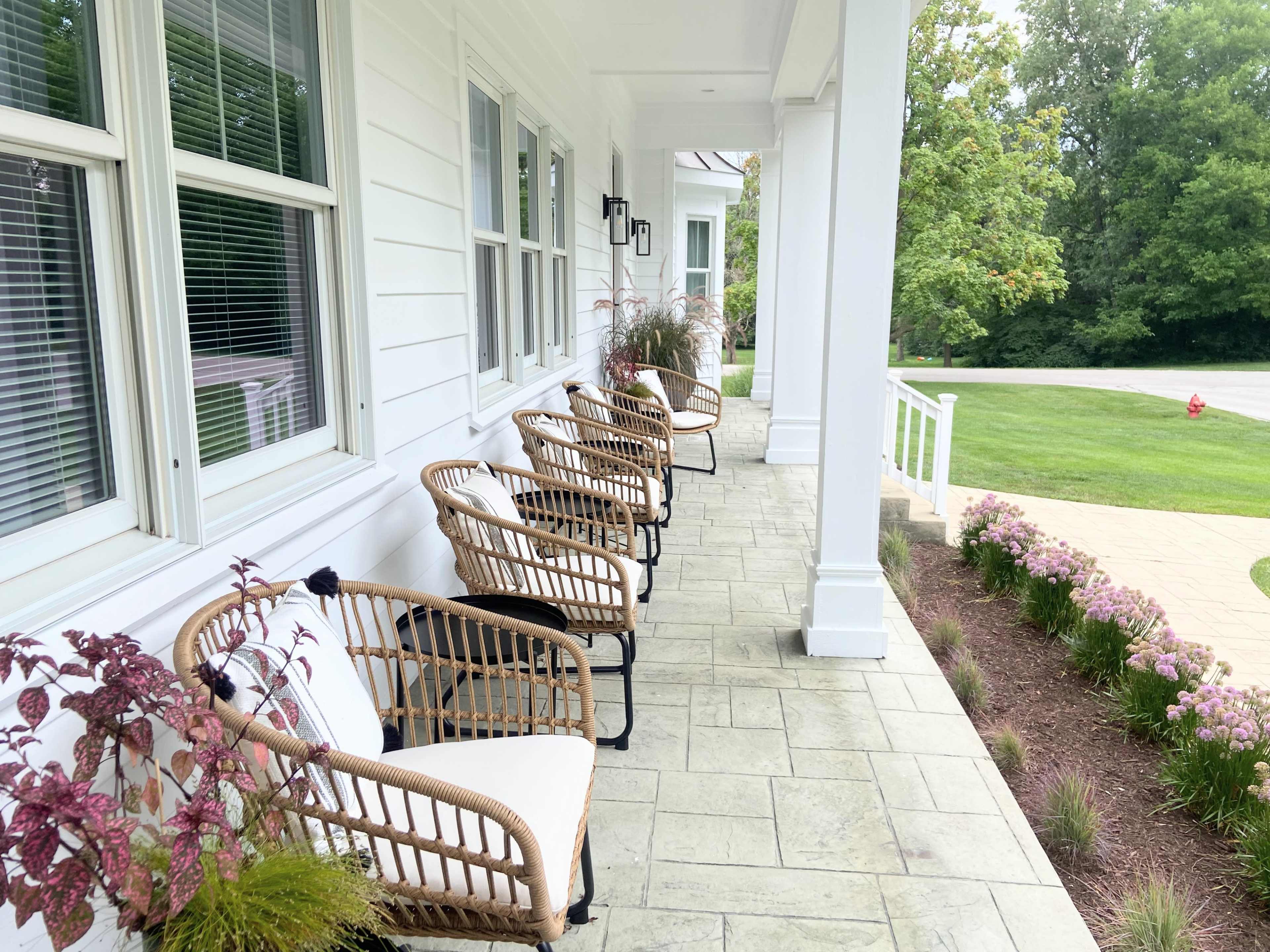 A row of wicker chairs is positioned on a covered porch beside potted plants and a grassy lawn.