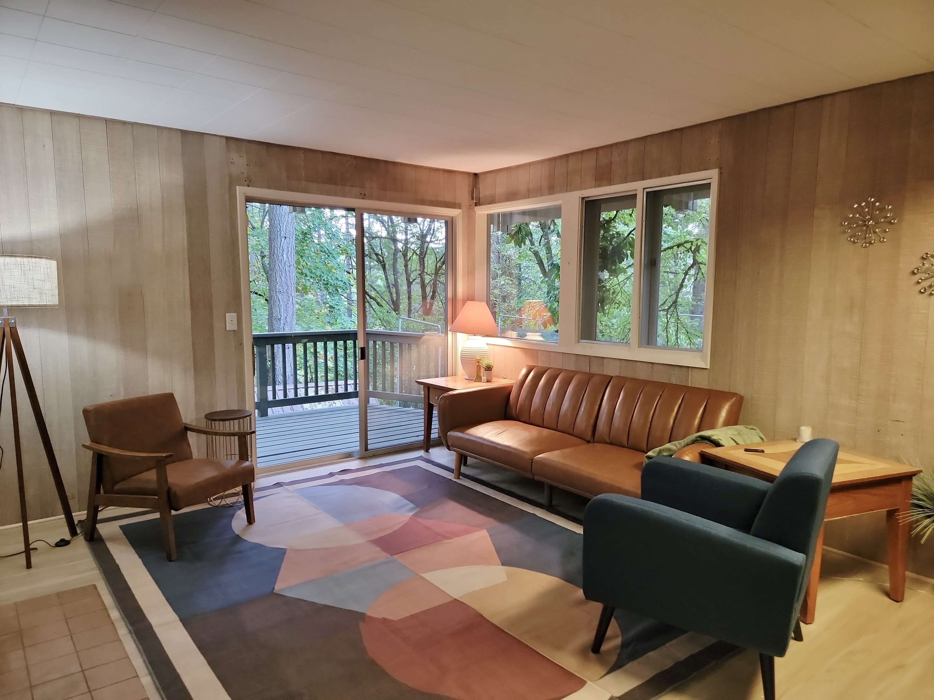 The image shows a living room with a brown leather sofa, a blue armchair, a wooden side table, and large windows overlooking a forested view.