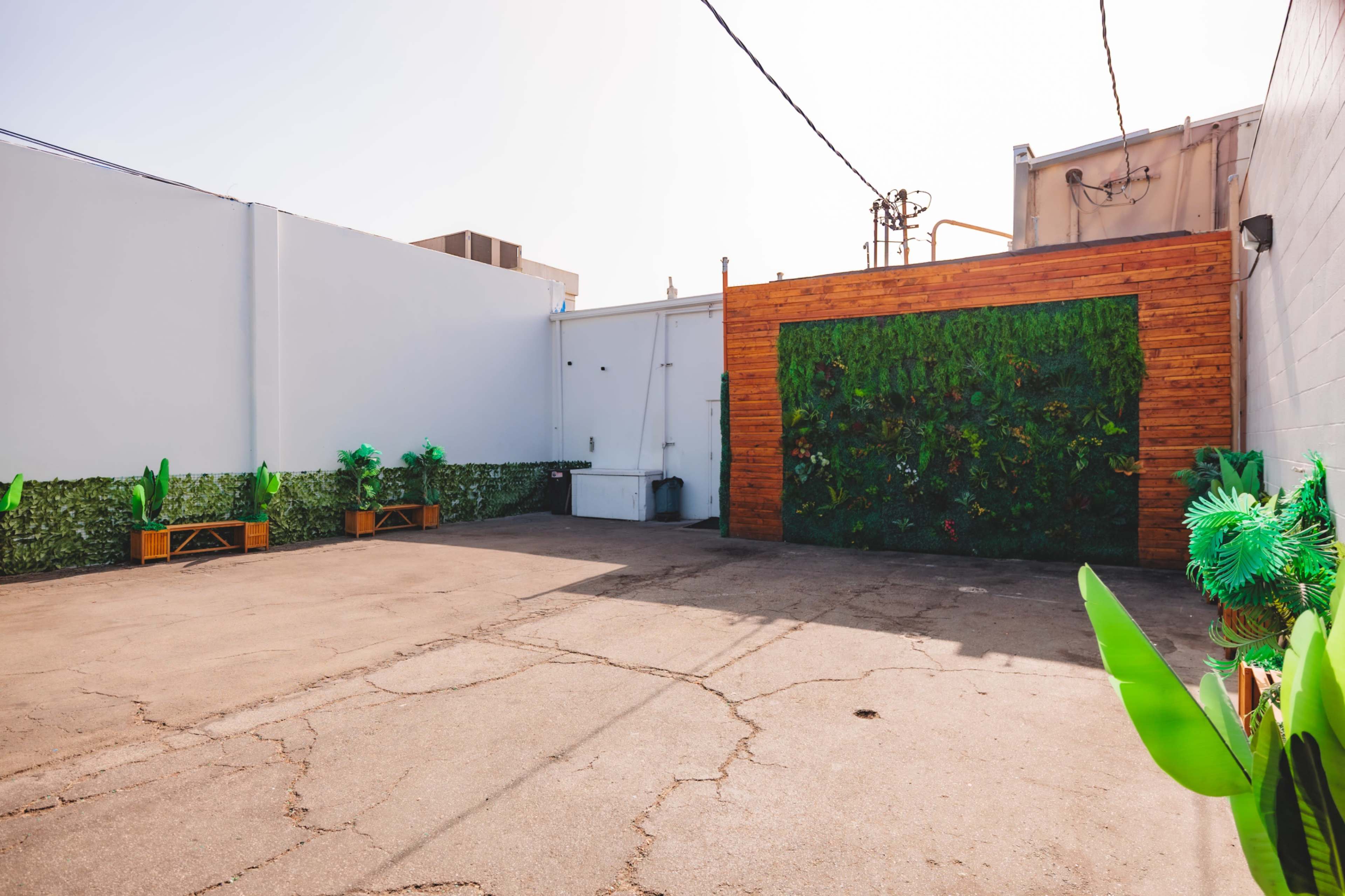 The image shows an outdoor space with a concrete floor, a green wall feature made of plants, and wooden accents, surrounded by artificial greenery decorations.