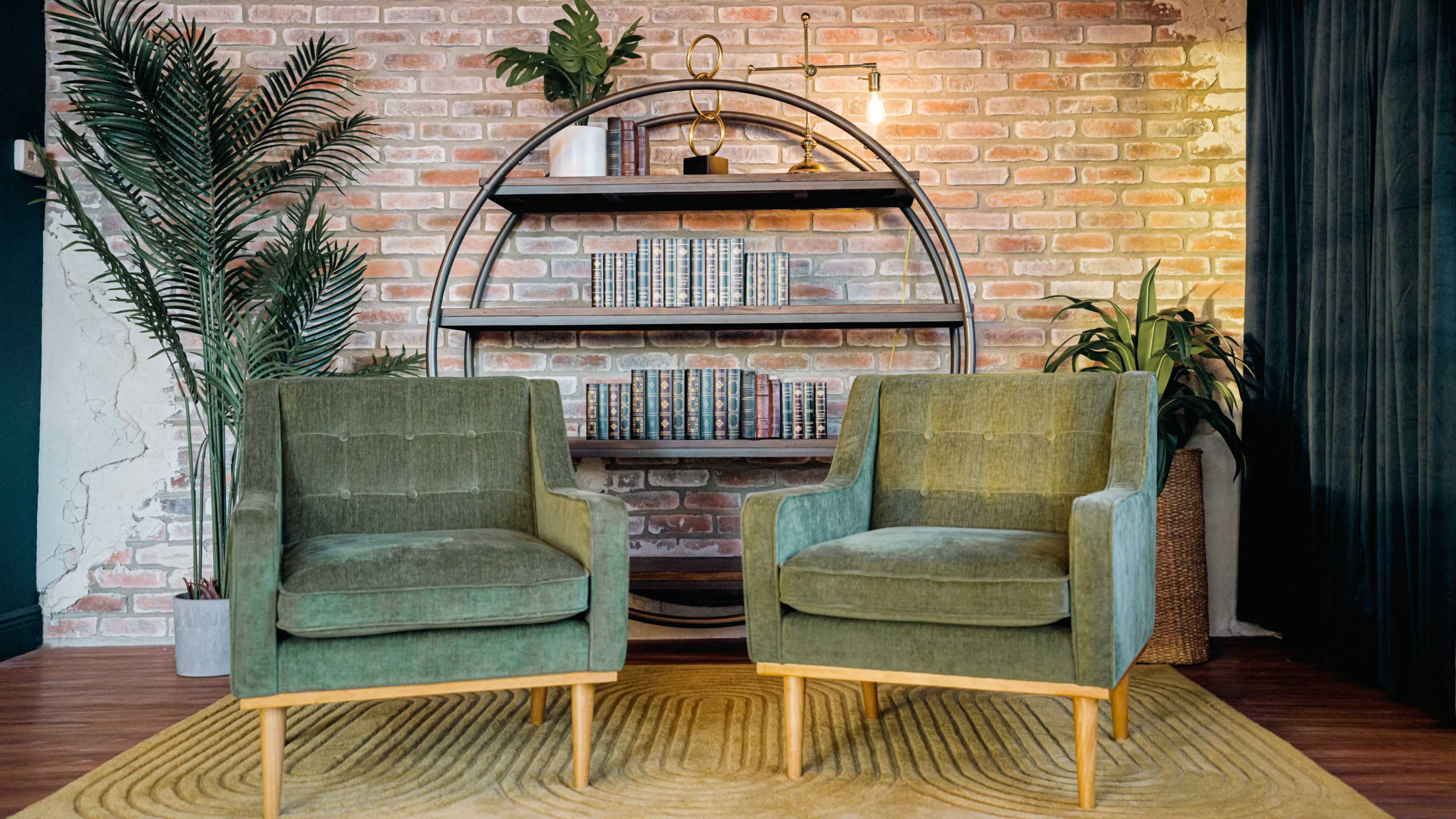 The image shows two green chairs positioned in front of a round shelf holding books, set against a brick wall with decorative plants.