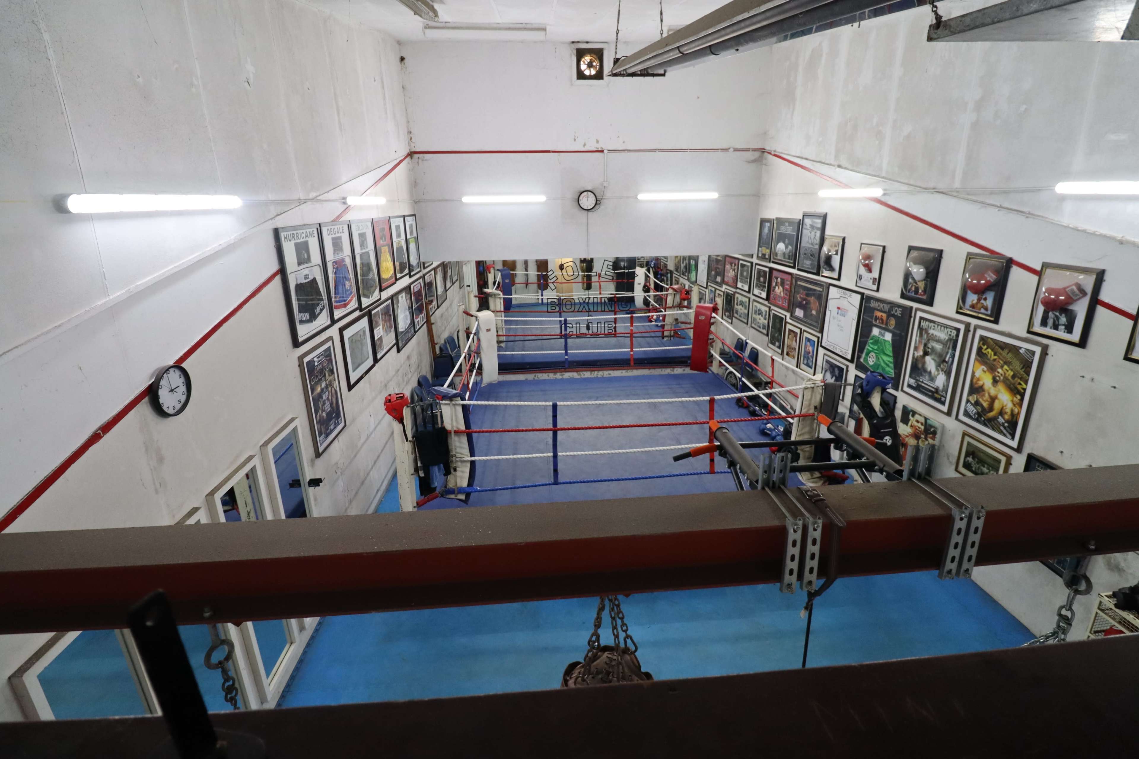 The image shows an indoor boxing gym with red and blue boxing rings, surrounded by framed photographs on the walls.