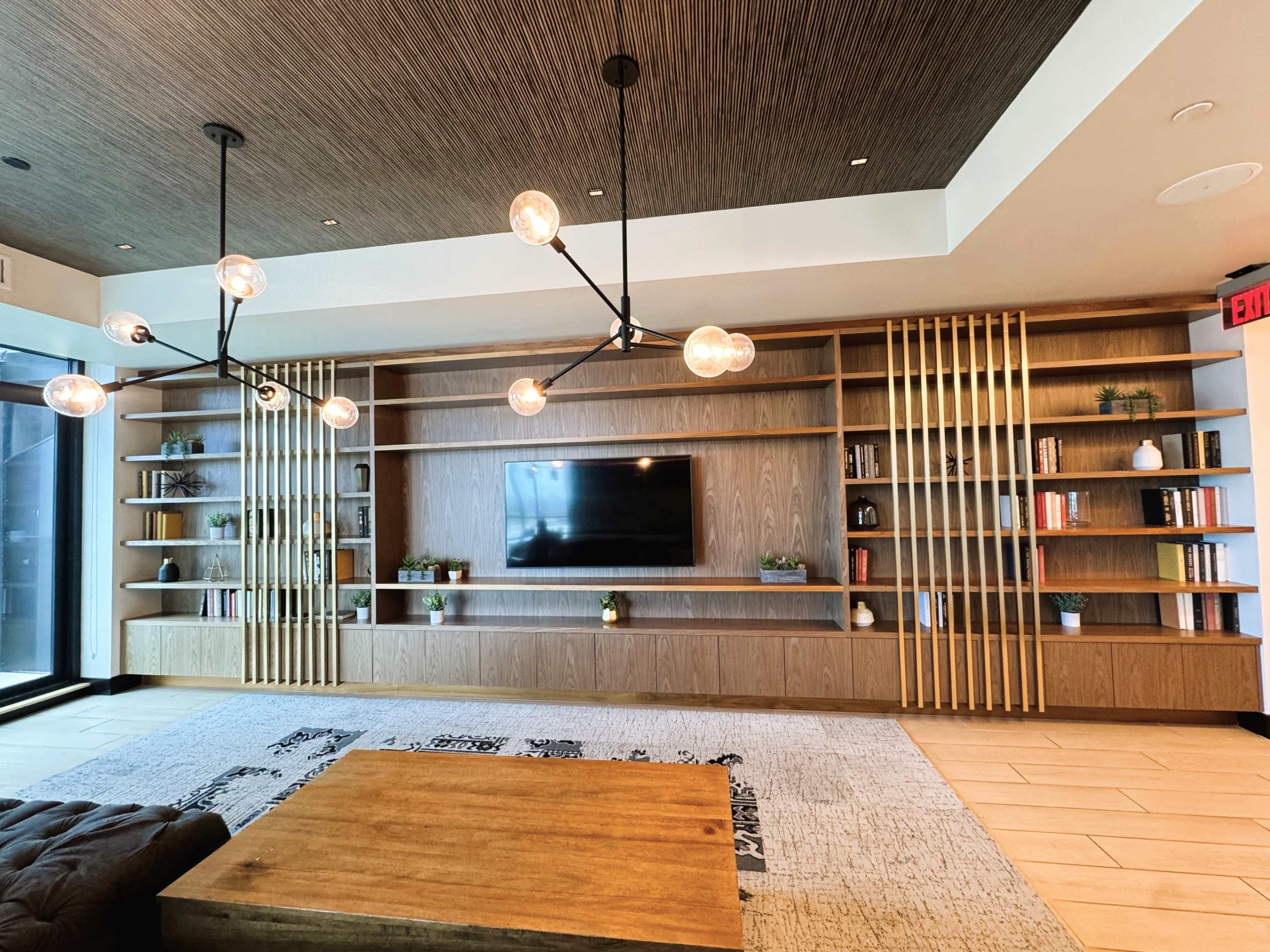 The image shows a modern lounge area with a wooden shelving unit, a large television mounted on the wall, and a low coffee table on a textured rug.