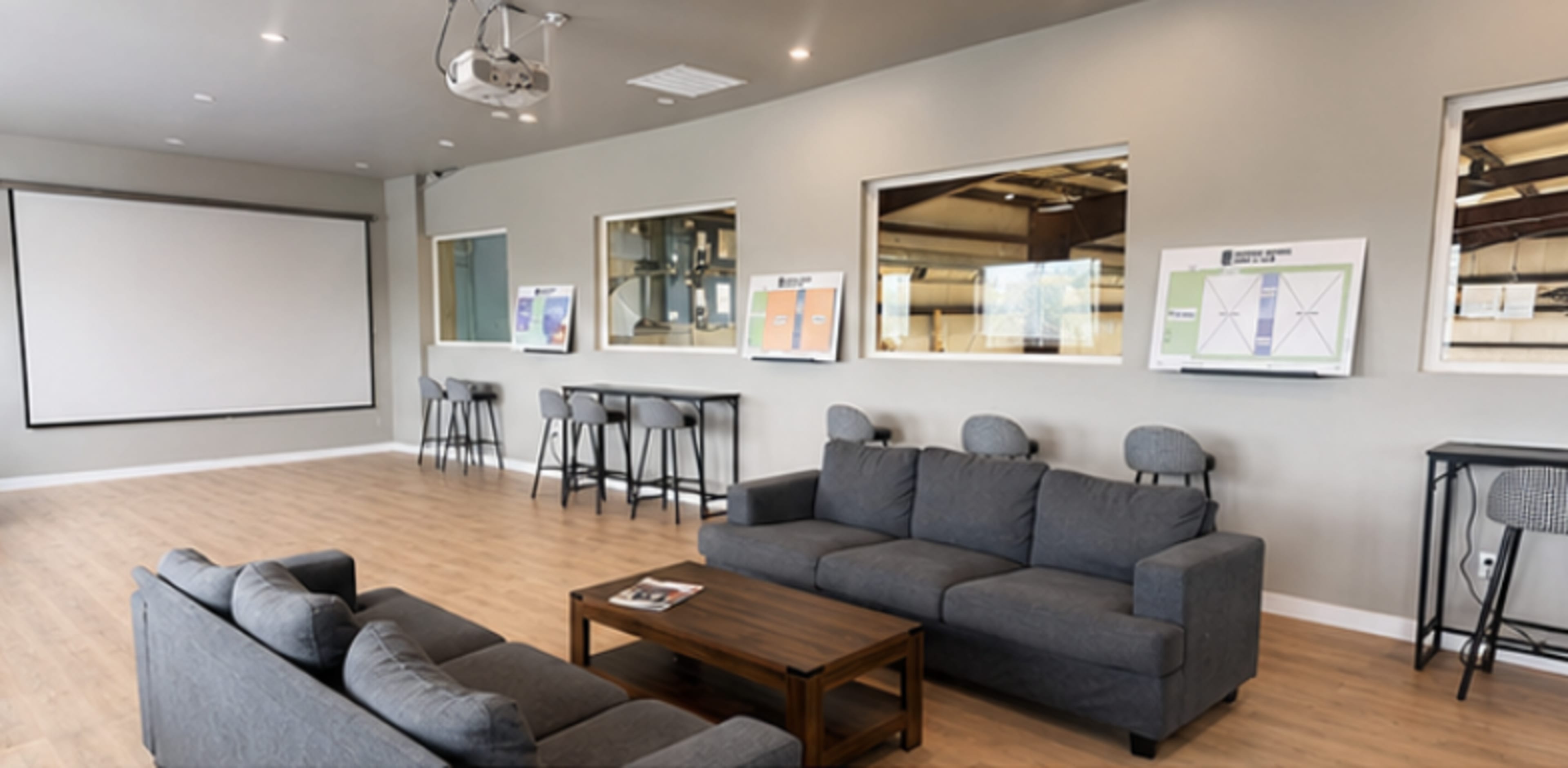 The image shows a modern room with a projection screen, grey sofas arranged around a wooden coffee table, and informational displays on the walls.