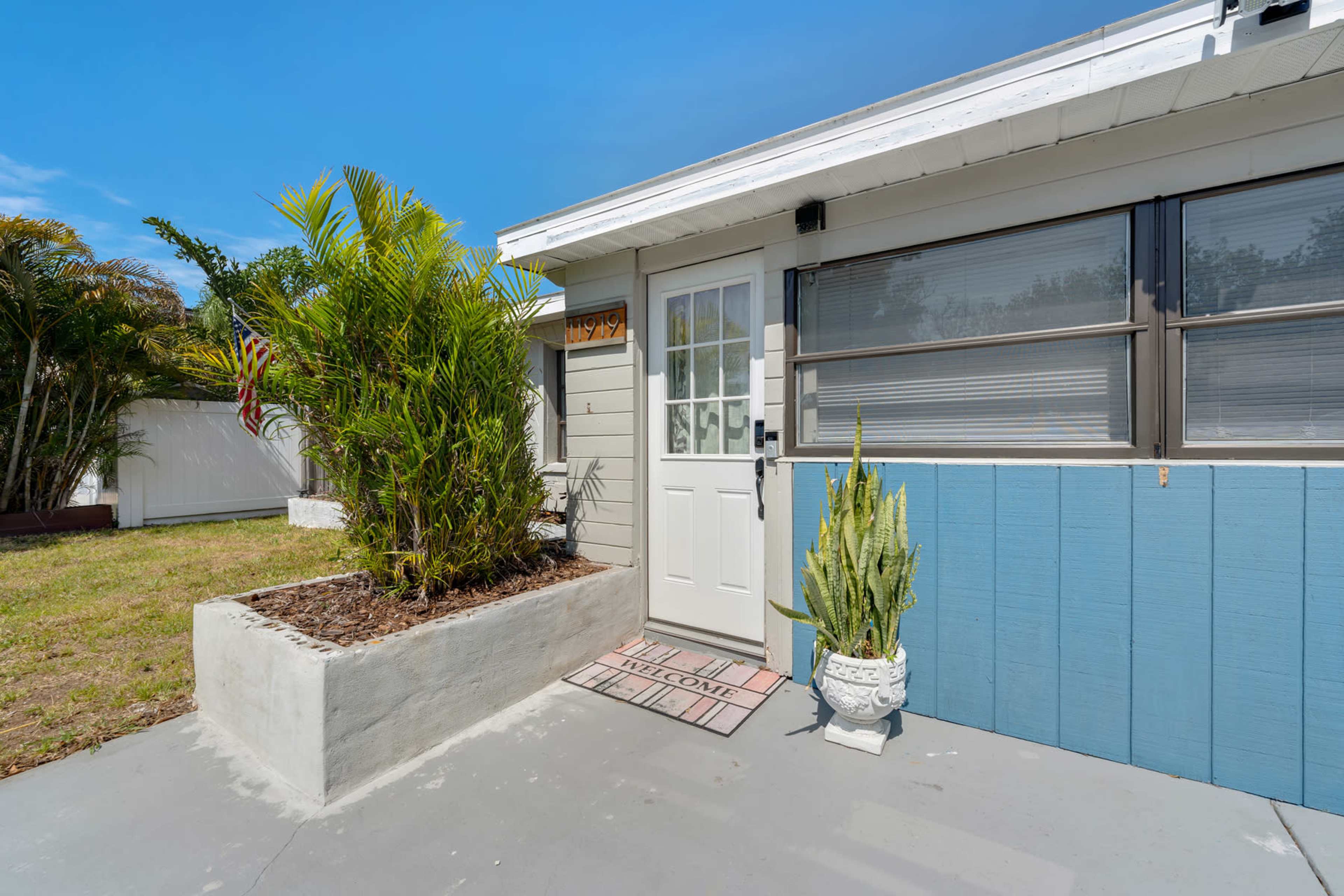 A small entrance area features a door with a welcome mat, surrounded by plants and a landscaped space.