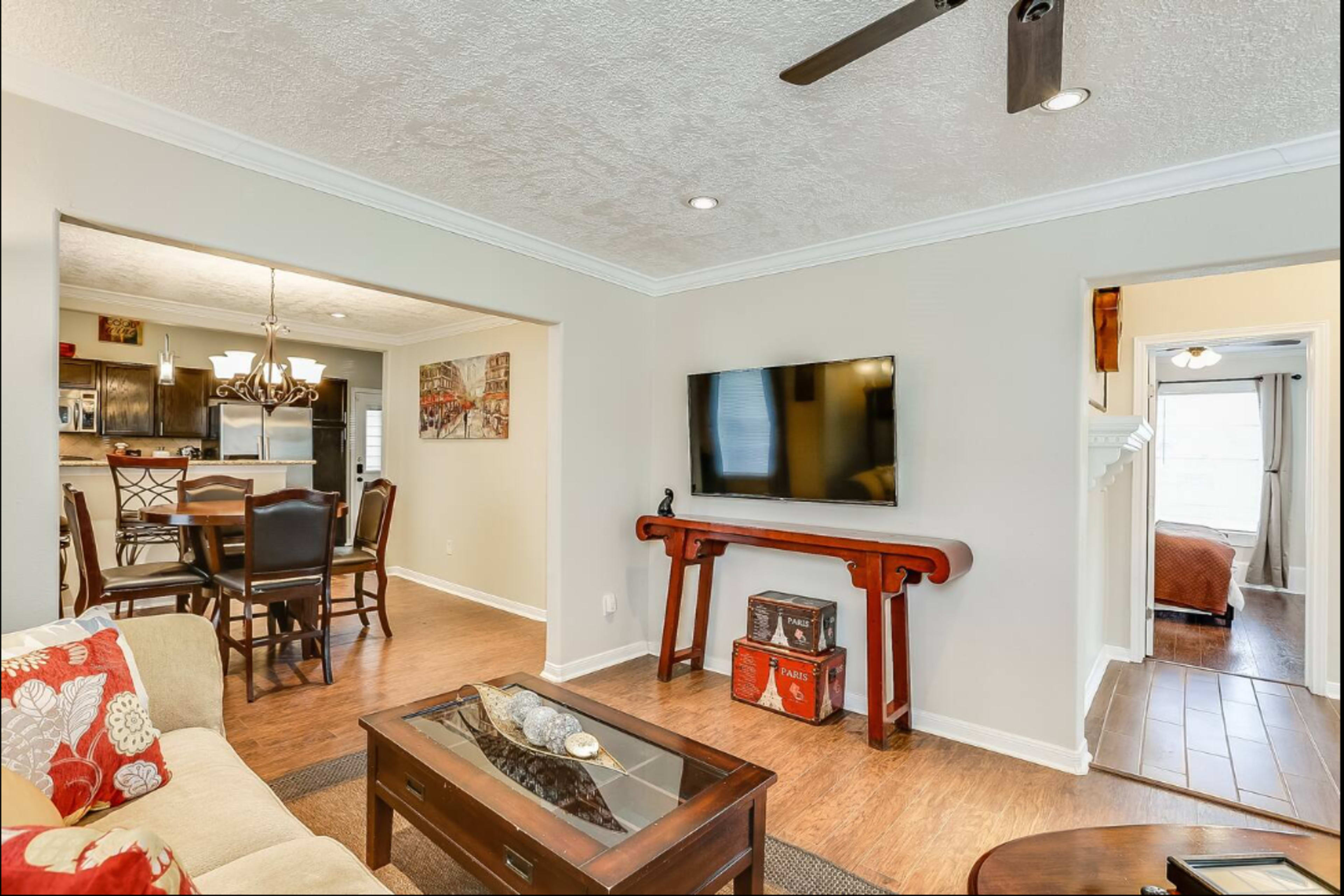The living room features a beige sofa with red accents, a wooden coffee table, and a mounted TV on the wall, leading into a dining area and visible bedroom.