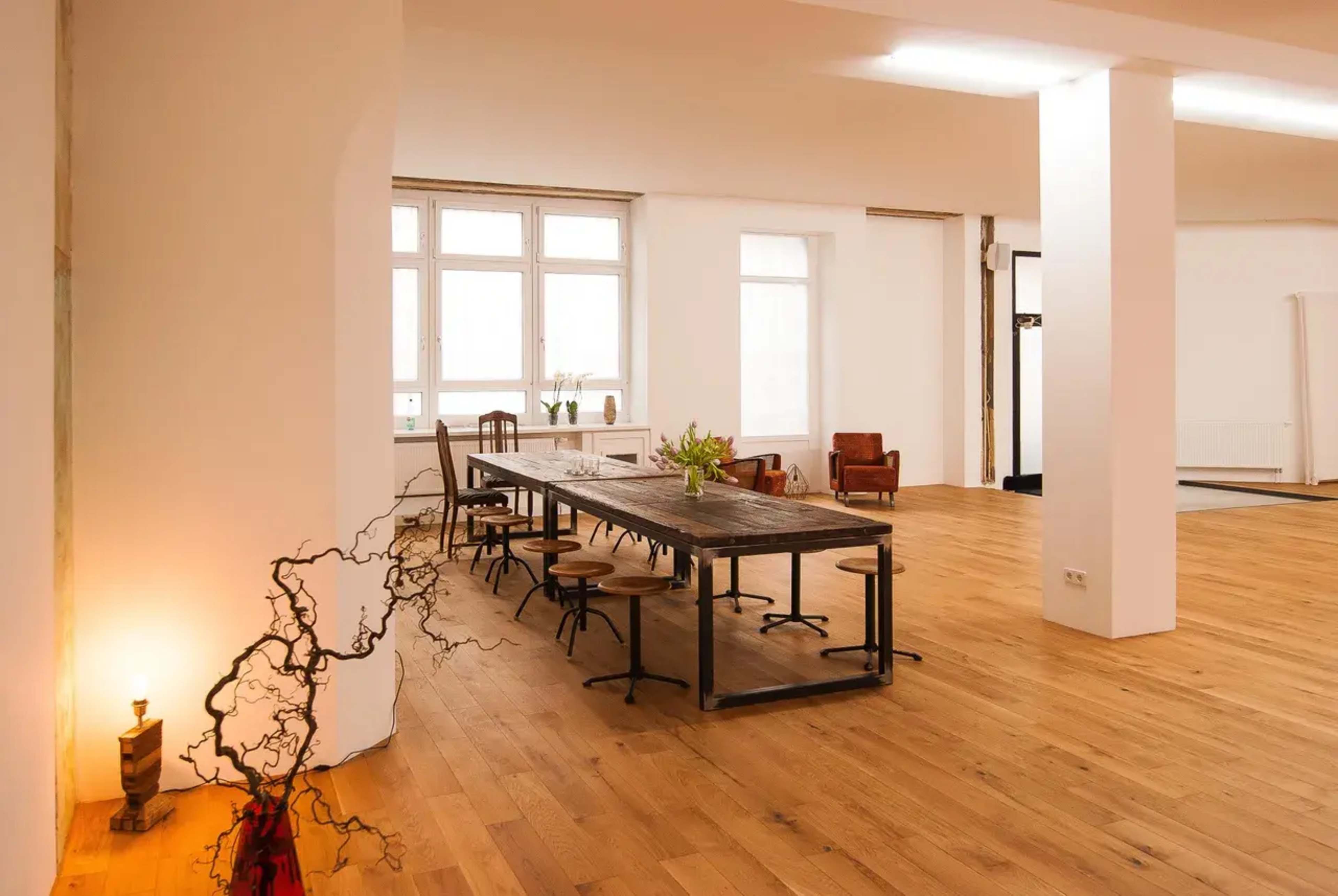 The image shows a spacious, well-lit room with a long wooden dining table, surrounded by chairs, and featuring wooden flooring and large windows.